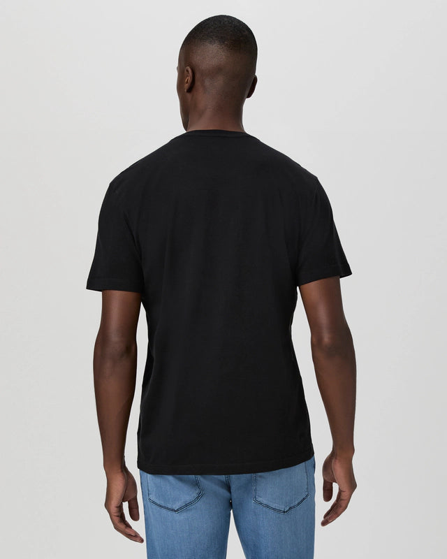 A man stands with his back to the camera, wearing a Paige Black Cotton Sorden Crew Neck T-Shirt and light blue jeans. His arms rest at his sides against a plain, light gray background. , available at Richard Gelding Mayfair