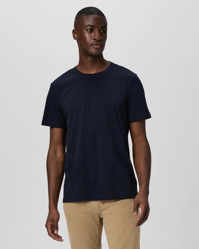 A man stands against a light background wearing the Paige Deep Anchor Navy Cotton Sorden Crew Neck and light beige pants. He looks slightly to the side with a relaxed expression, right arm at his side and left hand slightly raised. , available at Richard Gelding Mayfair