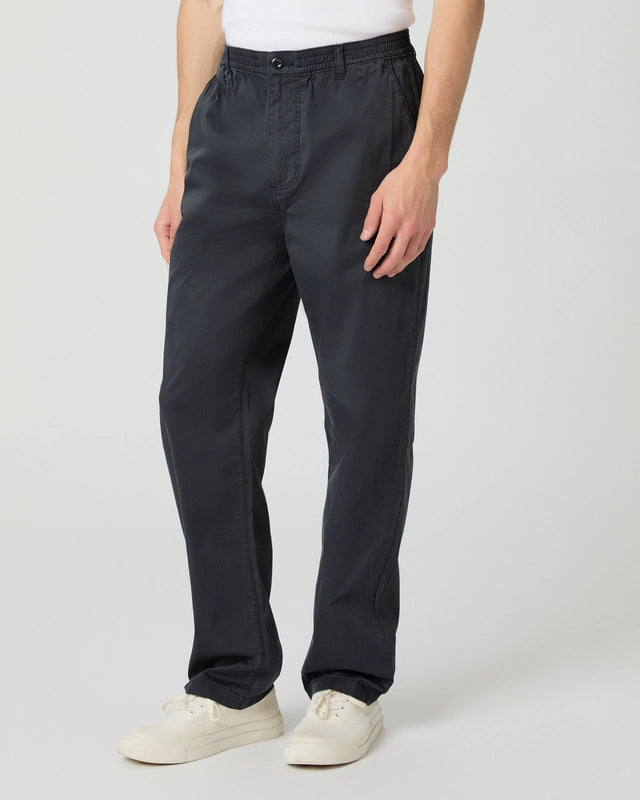 A person stands against a plain light background, wearing a white T-shirt tucked into Paige Navy Cotton Stretch 'Eberton' Trousers with an elastic waistband and relaxed fit, paired with white sneakers. Only the torso and legs are visible. , available at Richard Gelding Mayfair