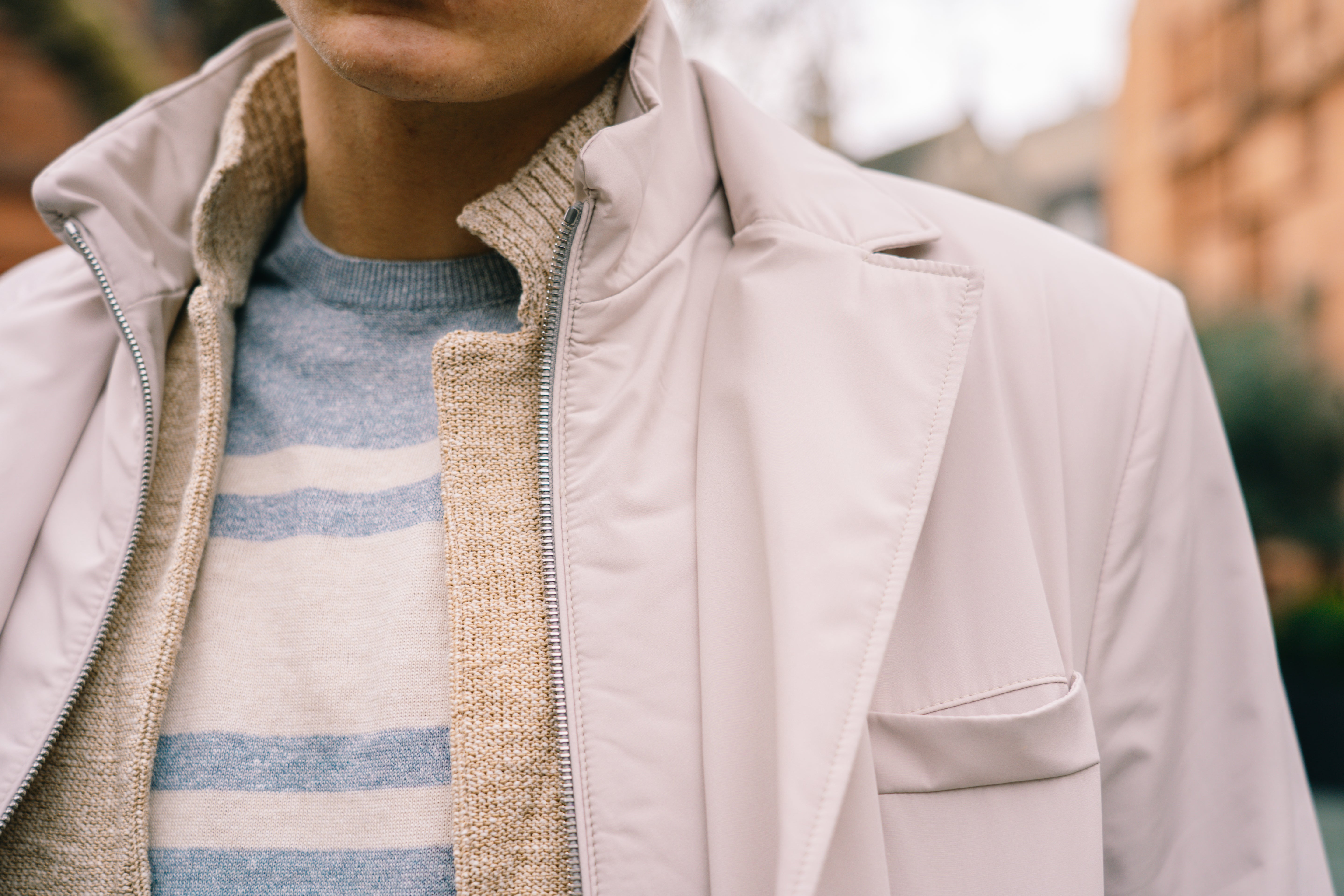 A person wears a beige coat over the Gran Sasso Grey Stripe Cotton/Linen Knitted T shirt. The image is cropped to the lower face and upper body, with blurred buildings and greenery in the background. , available at Richard Gelding Mayfair
