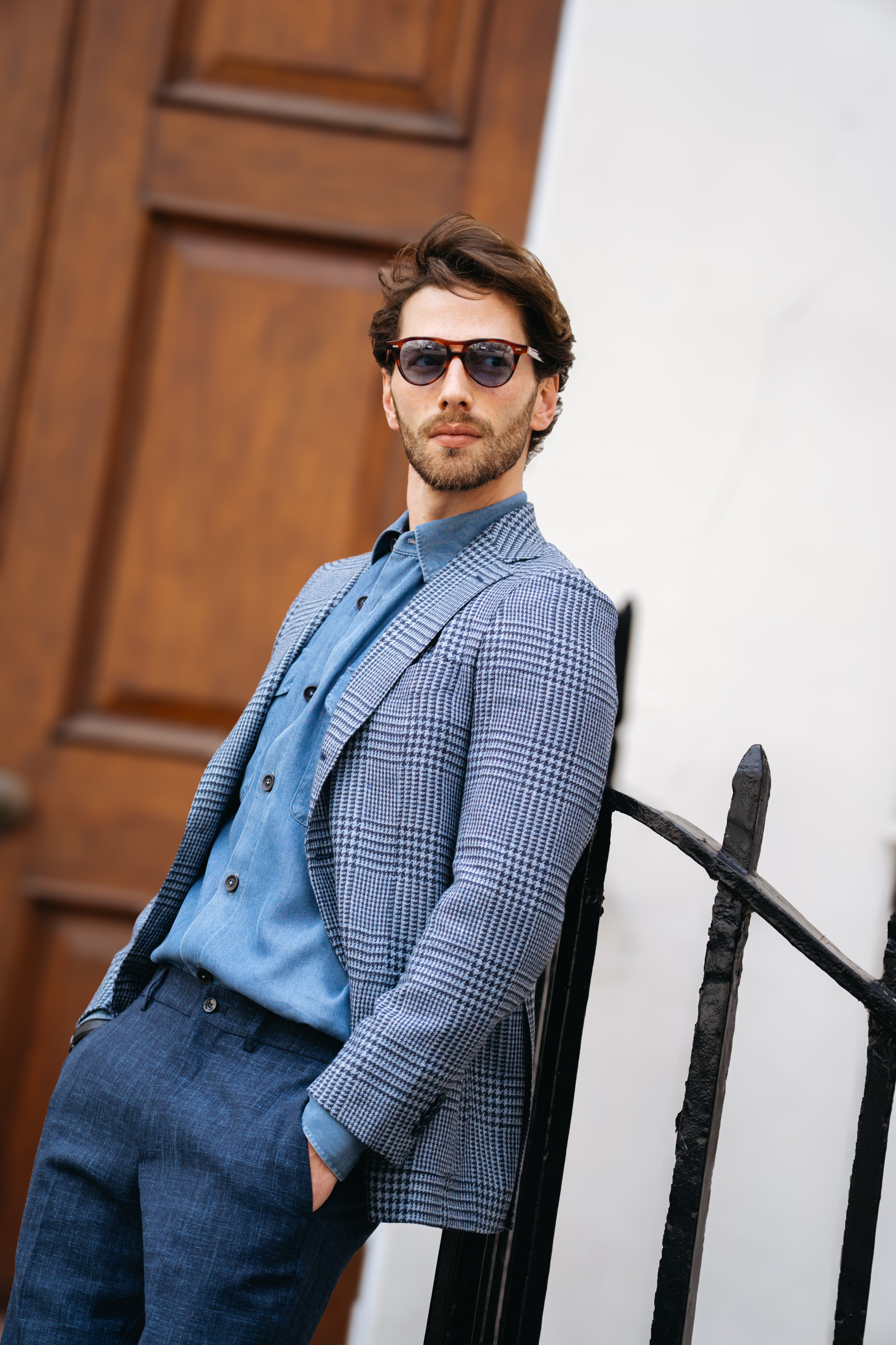 A man with brown hair and a beard, wearing sunglasses, a blue shirt, and the Cavour Blue Glencheck Wool/Silk/Linen Blazer, stands outdoors leaning against a black metal fence. A large wooden door and white wall are in the background. , available at Richard Gelding Mayfair