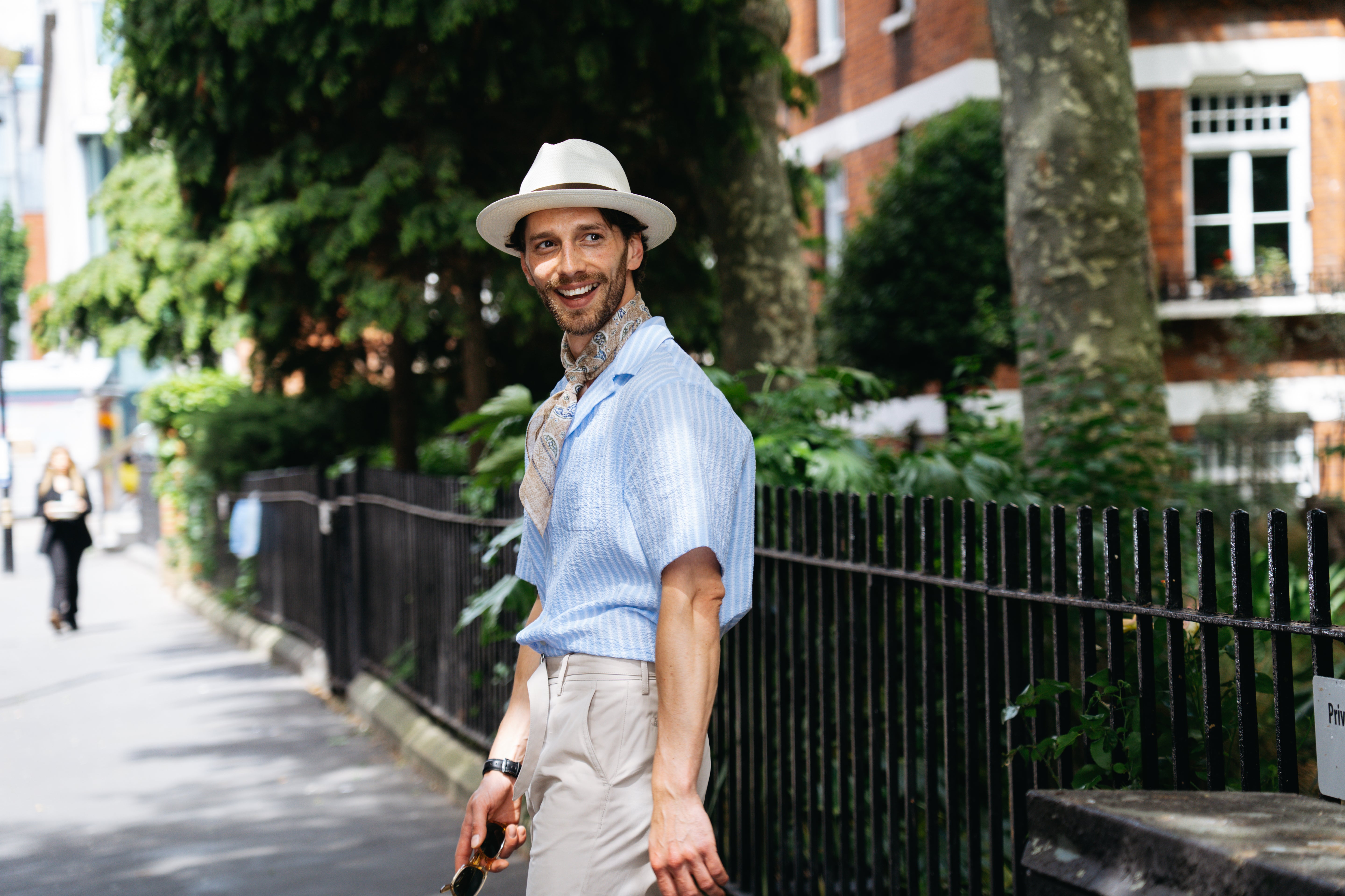 A smiling man walks along a sunlit, tree-lined city street in an Altea Baby Blue Cotton Seersucker Cuban Shirt, beige trousers, and white hat, holding sunglasses. Brick buildings and a black iron fence line the leafy background. , available at Richard Gelding Mayfair