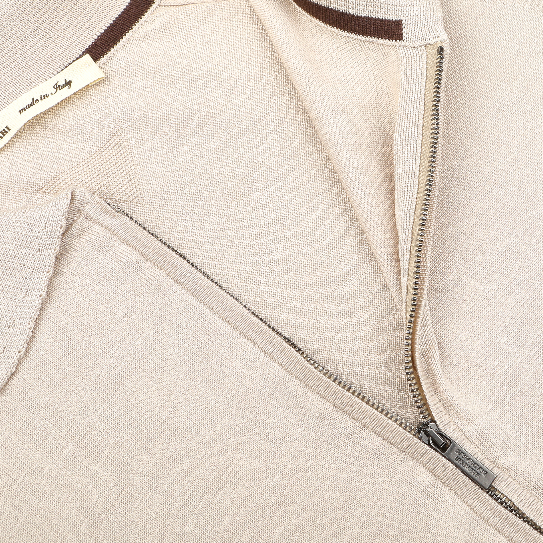Close-up of a beige silk cotton full zip polo shirt by Maurizio Baldassari, partially open zipper shows a brown and white "made in Italy" tag. This exquisite piece embodies the essence of Italian knitwear, merging contemporary design with timeless elegance.