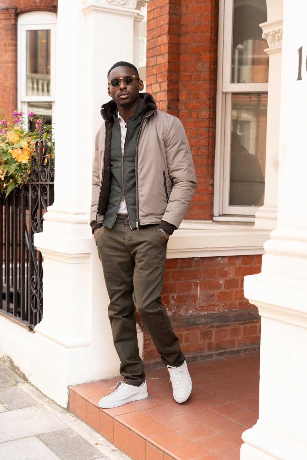 A man stands on a brick porch outside a building with white pillars. He wears sunglasses, a beige jacket over a green hoodie and shirt, olive pants, and white sneakers. His hands are in his pockets and he faces the camera confidently. , available at Richard Gelding Mayfair