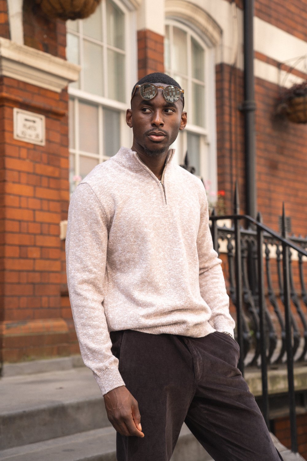A man stands outdoors in front of a brick building with white-framed windows. He wears a light beige knit pullover, dark trousers, and sunglasses resting on his head. His hands are relaxed at his sides, and he looks to the left with a thoughtful expression. , available at Richard Gelding Mayfair