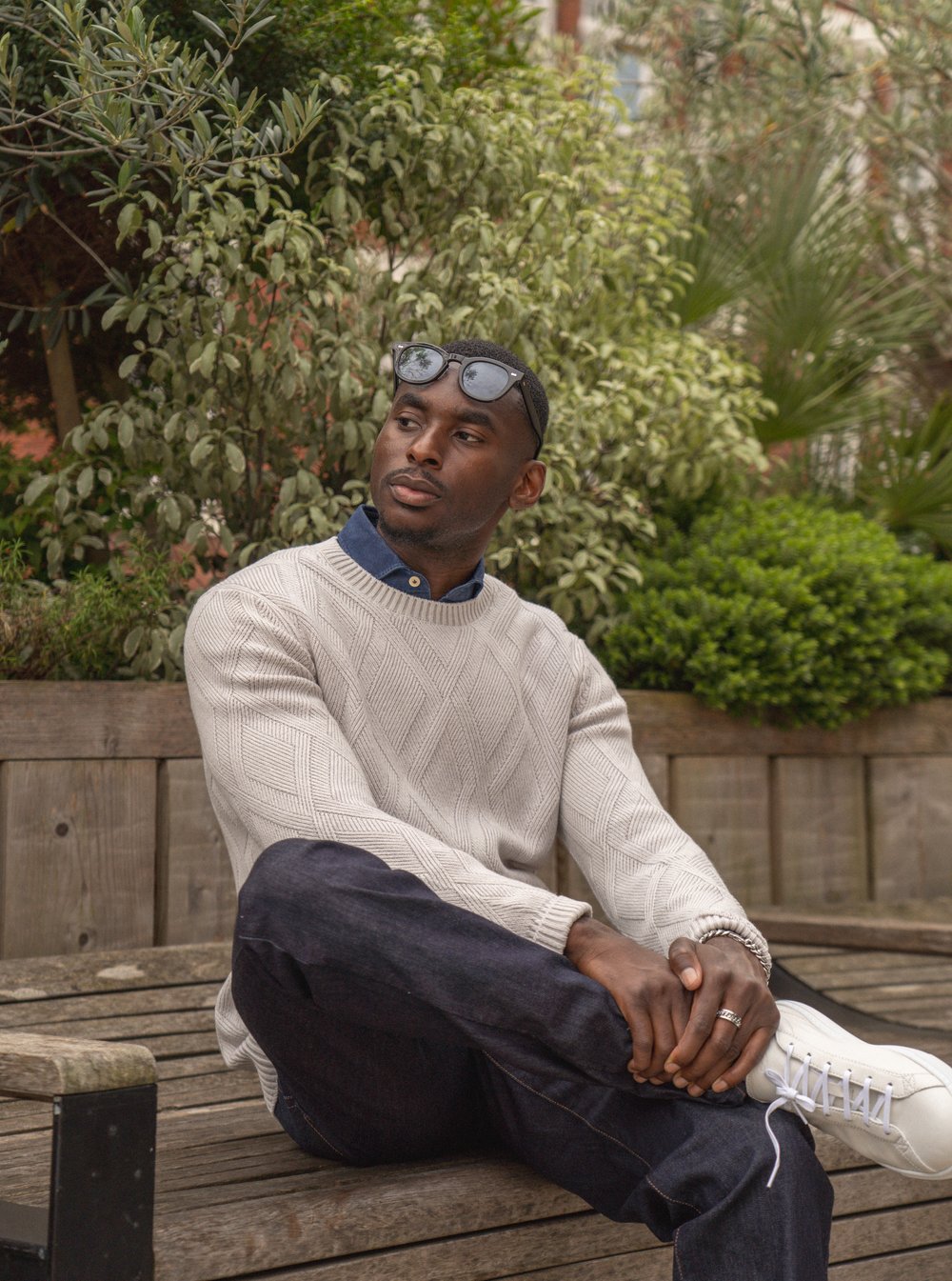 A man sits on a wooden bench outdoors, surrounded by green leafy plants. He wears a cream cable-knit sweater over a blue collared shirt, dark jeans, white sneakers, and sunglasses on his head. He looks to the side with a relaxed expression, hands clasped. , available at Richard Gelding Mayfair