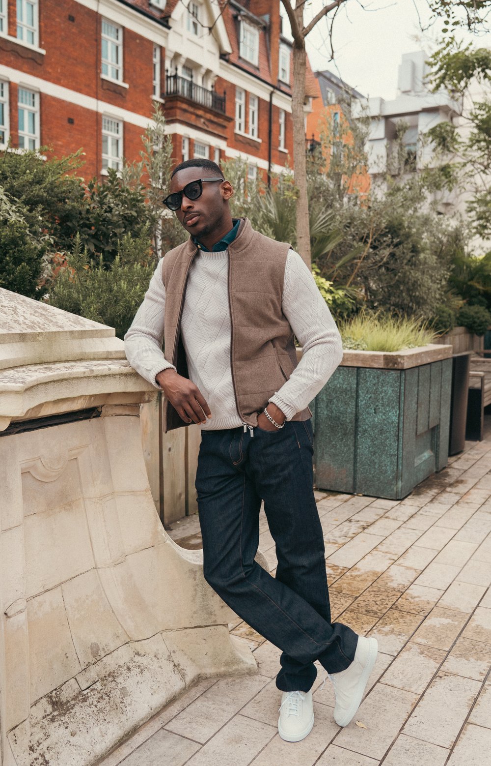 A stylish man wearing a beige sweater, brown vest, dark jeans, white sneakers, sunglasses, and a cap leans casually on a stone railing in an urban outdoor setting with greenery and red brick buildings in the background. , available at Richard Gelding Mayfair