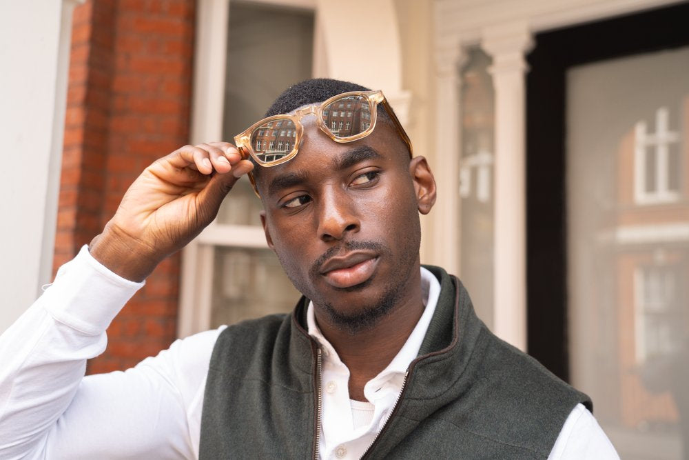 A man with short hair lifts his sunglasses while looking to the side. He wears a white shirt and a dark vest. The background shows red brick and white pillars, indicating an urban or residential setting. , available at Richard Gelding Mayfair
