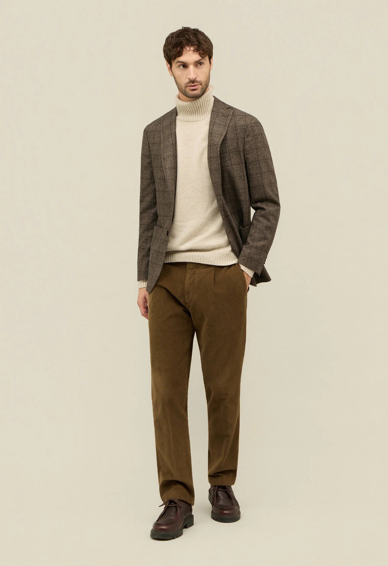 Man wearing a brown blazer, beige sweater, and brown pants on a beige background