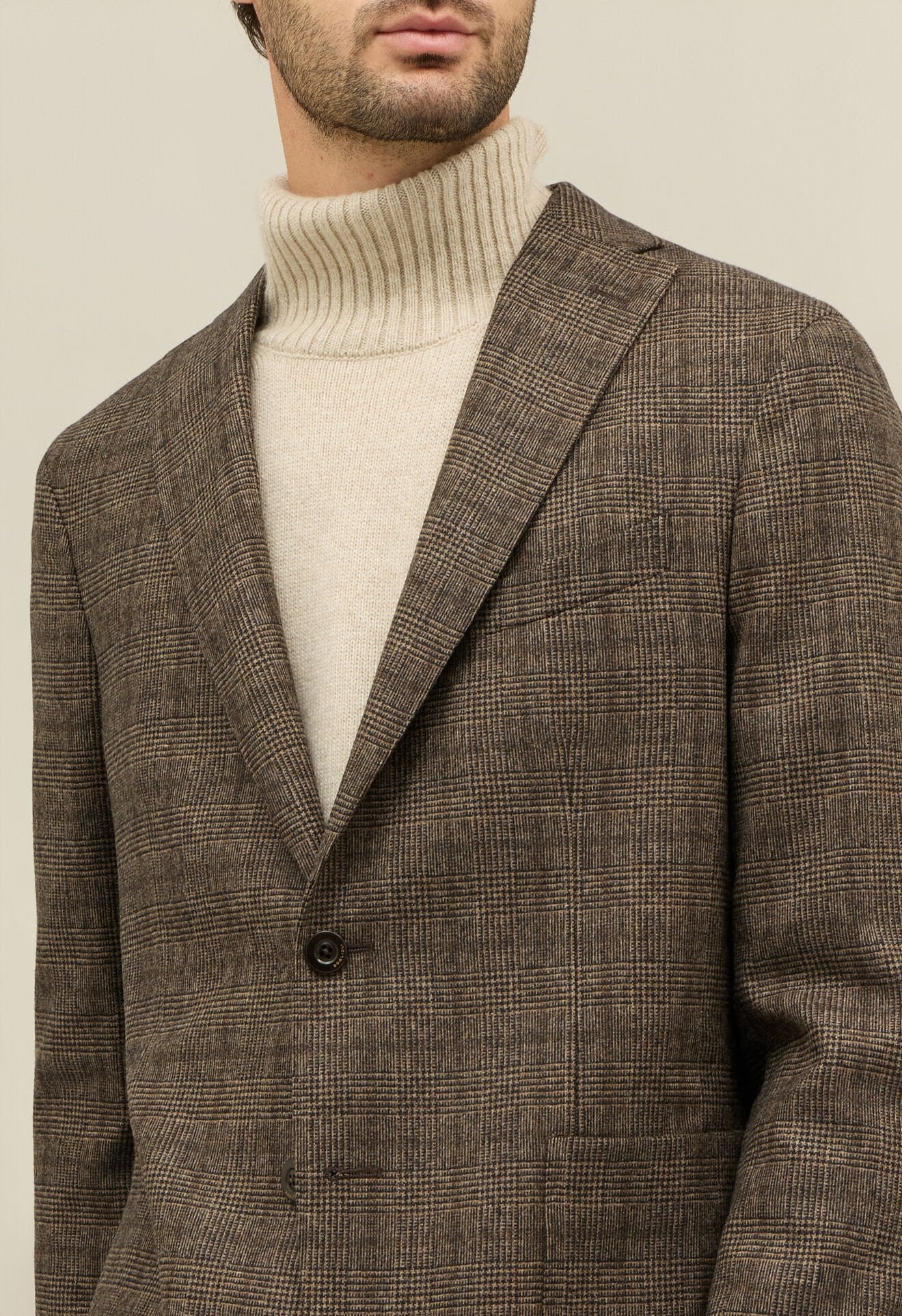 A man wears a beige ribbed turtleneck under the Boglioli Brown Prince Of Wales Check Linen Jacket. With only his mouth and chin visible, a plain beige background accentuates his outfit’s textures and check pattern. , available at Richard Gelding Mayfair