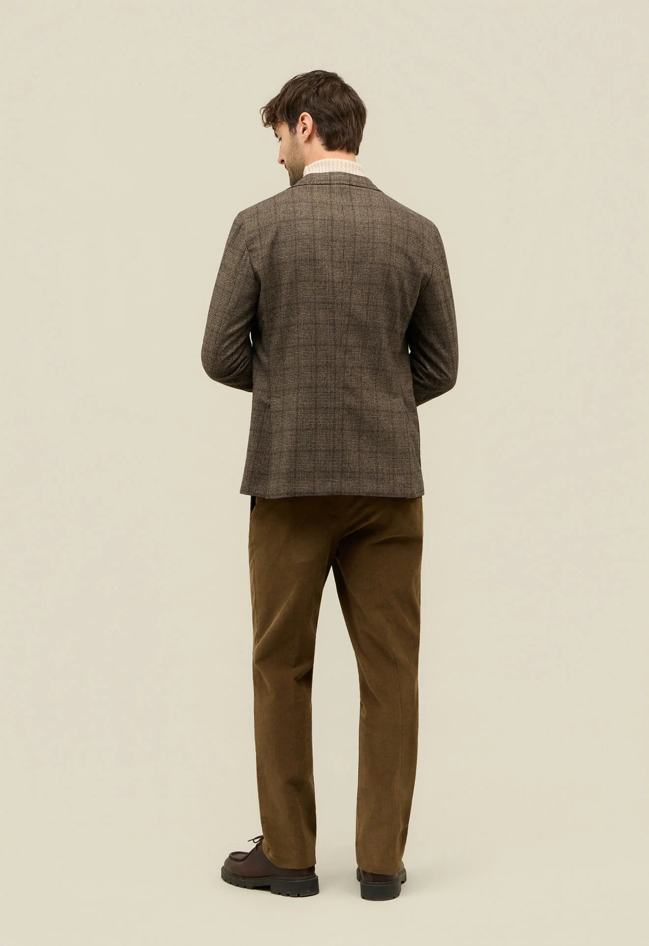 A man stands with his back to the camera against a beige background, wearing a Boglioli Brown Prince Of Wales Check Linen Jacket, white shirt, brown corduroy pants, and dark brown shoes. His short brown hair and relaxed posture finish the look. , available at Richard Gelding Mayfair