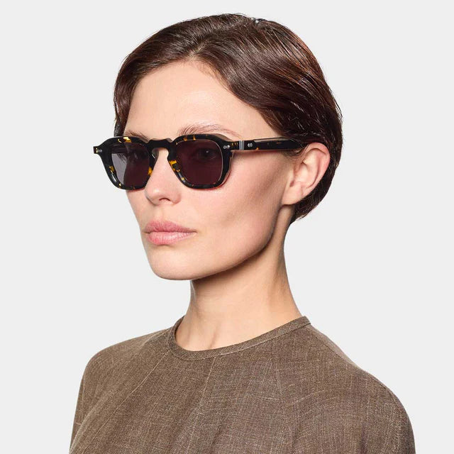 A woman with short brown hair wears TBD Eyewear's Ply Black Havana | Grey rectangular sunglasses, handcrafted in Italy, and a brown textured top. She faces slightly right against a plain light-gray background, her expression neutral. , available at Richard Gelding Mayfair