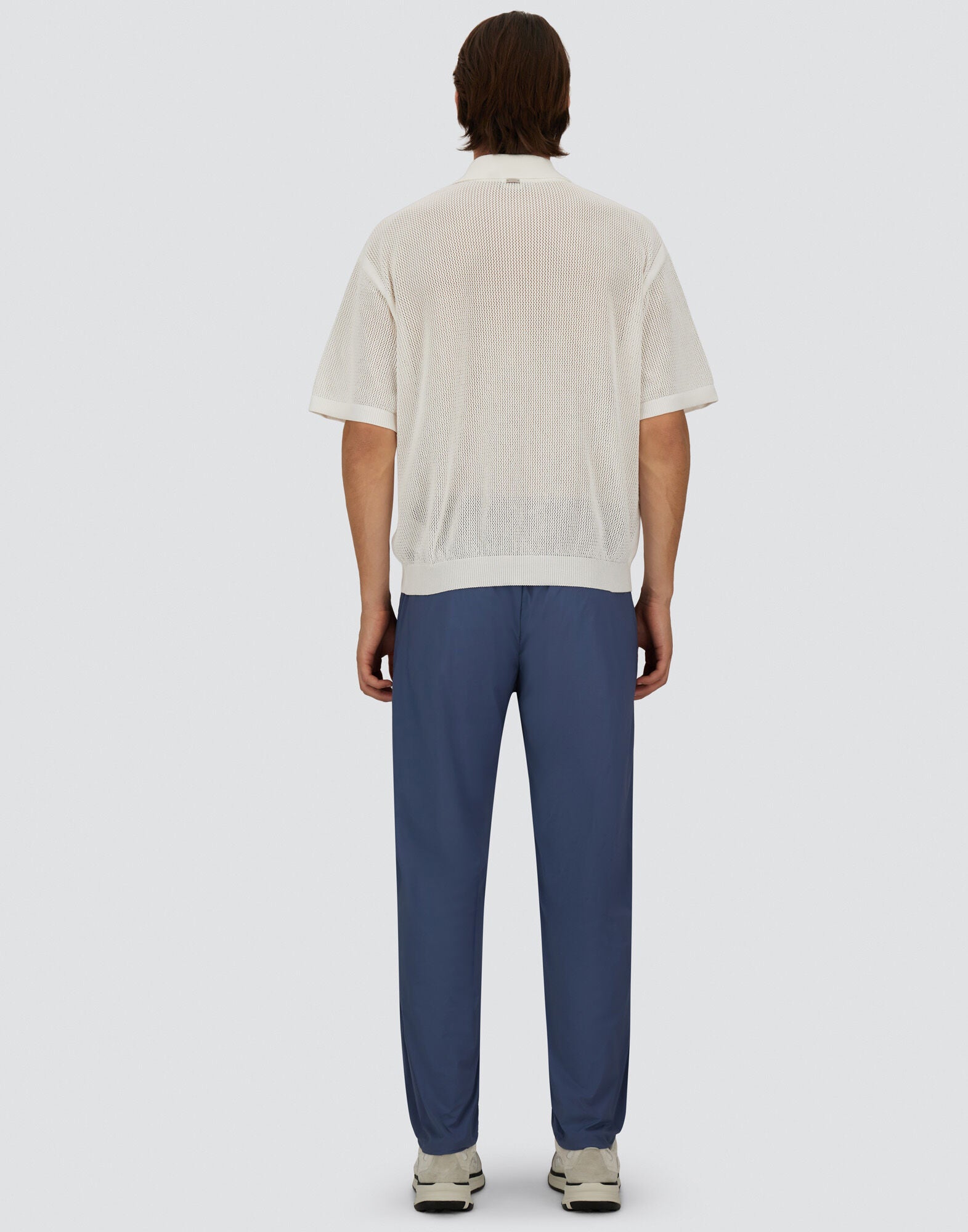 A man with medium-length brown hair stands facing away against a light-gray background, wearing Herno Blue Lightweight Nylon Drawstring Casual Trousers, a sheer white short-sleeve shirt, and white sneakers. His arms rest naturally by his sides. , available at Richard Gelding Mayfair