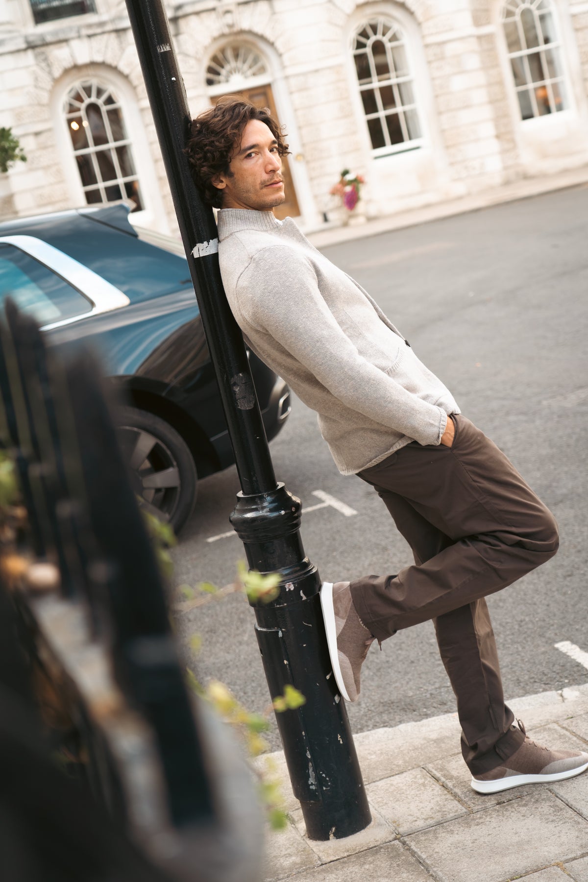 A man with curly hair, wearing the Maurizio Baldassari Beige Wool Cotton Winter Mouline Cortina Cardigan, brown pants, and sneakers, leans against a black lamppost on a city street. One leg is bent, hands in pockets. Behind him is a black car and a white stone building with arched windows. , available at Richard Gelding Mayfair