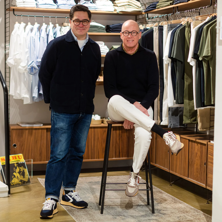 Two men in glasses pose in a clothing store. One stands in jeans and sneakers, smiling with hands behind back. The other, seated on a tall stool, wears white pants and beige sneakers. Shelves of folded shirts and hanging clothes fill the background. , available at Richard Gelding Mayfair