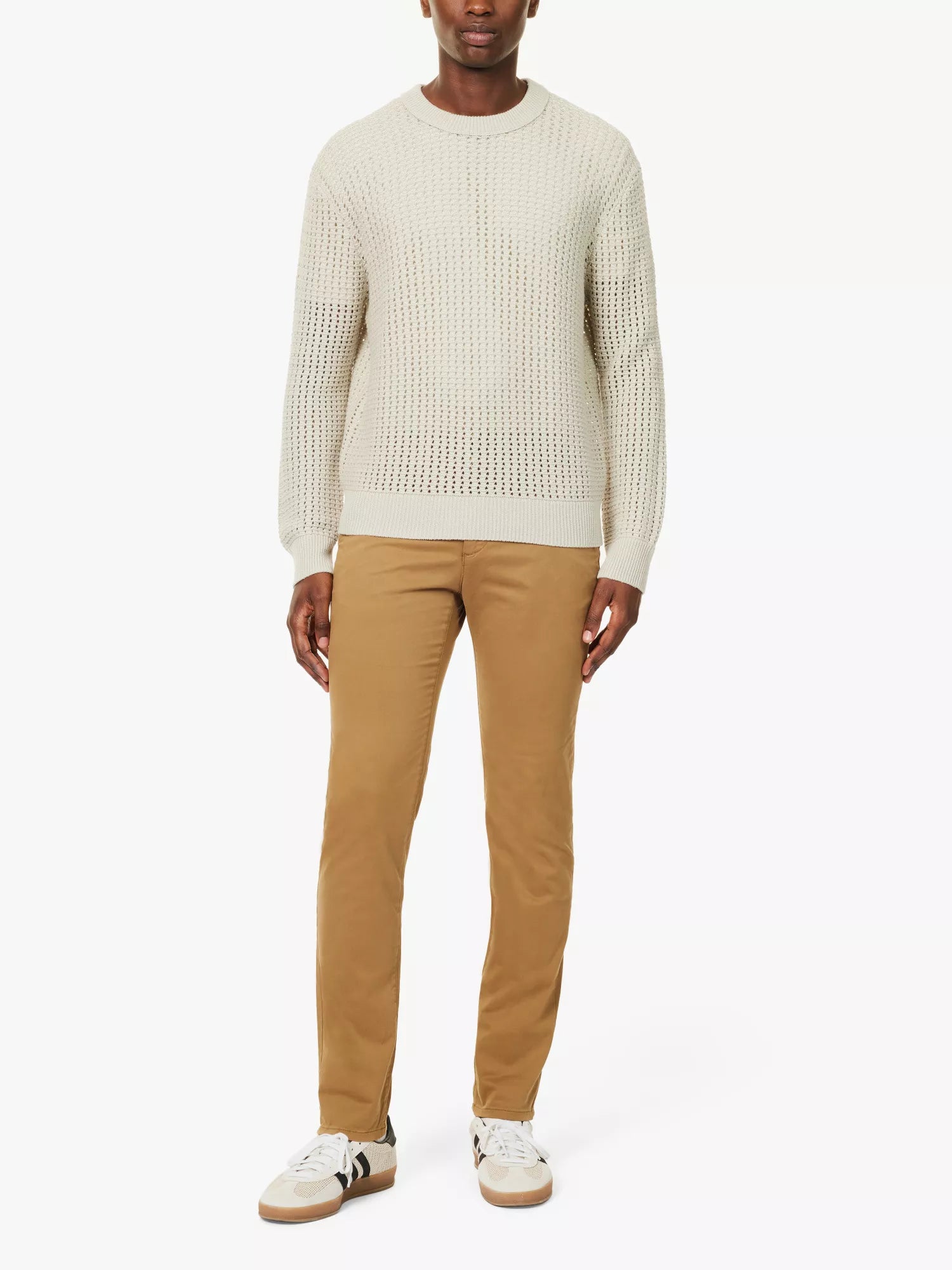 A person, head not visible, stands against a white background wearing a cream textured knit sweater, Paige Tobacco Brown Danford Cotton Chino pants, and white sneakers with beige soles and black accents. , available at Richard Gelding Mayfair