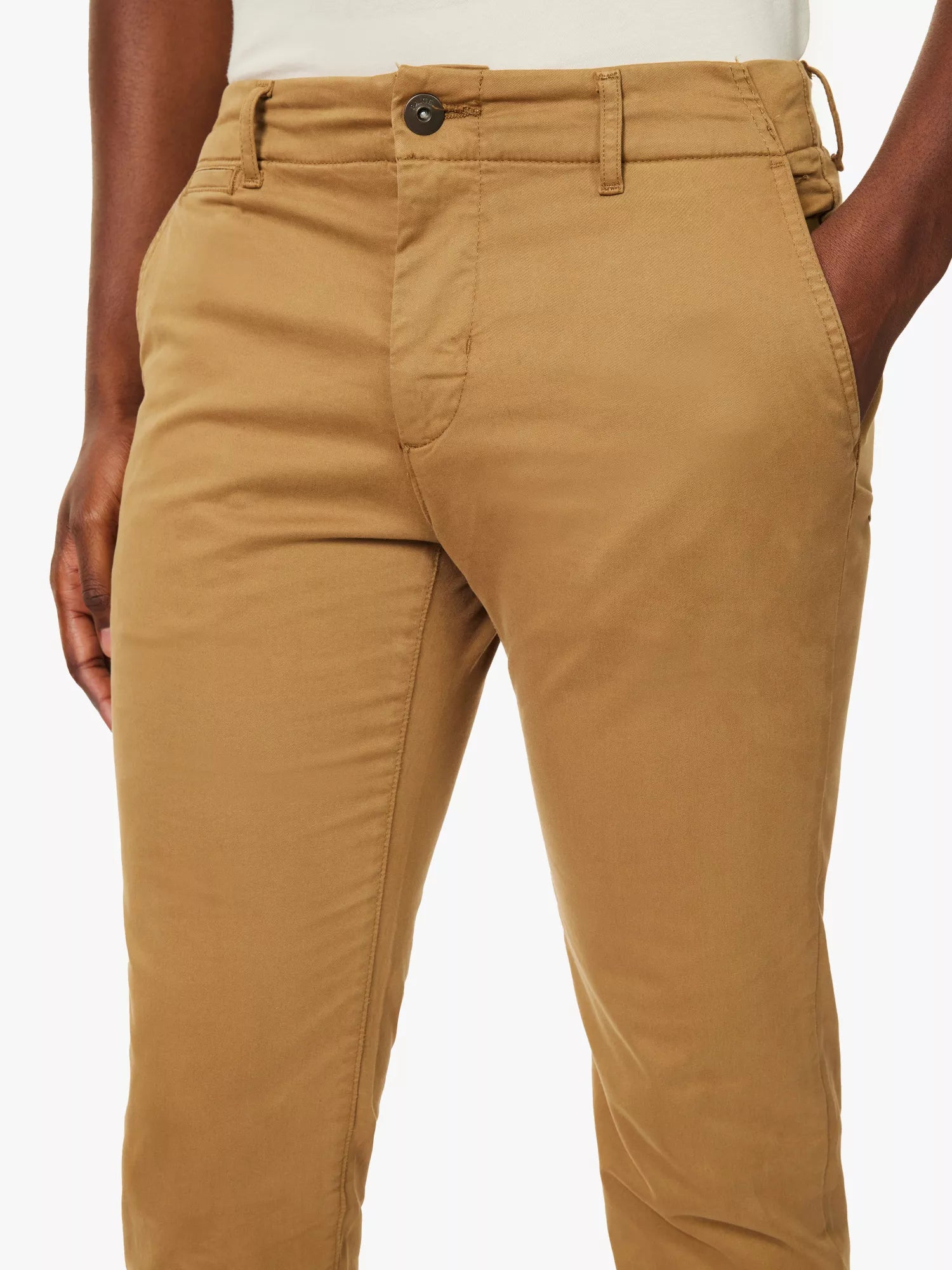 A person wears Paige's Tobacco Brown Danford Cotton Chino, featuring a button and zipper fly, belt loops, and front pockets. Their left hand rests partly in the pocket. Only the lower torso and upper legs are shown against a plain white background. , available at Richard Gelding Mayfair