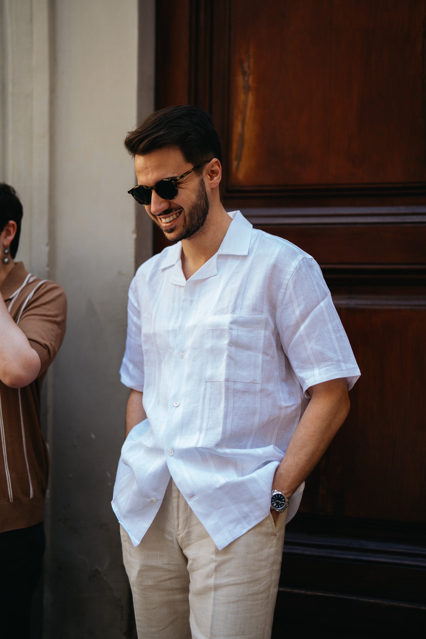 A man with dark hair and a beard, wearing sunglasses and a Maurizio Baldassari White Linen Short Sleeve Bowling Shirt with light-colored pants, stands smiling in front of a wooden door, hands in pockets. Another person is partly visible beside him. , available at Richard Gelding Mayfair