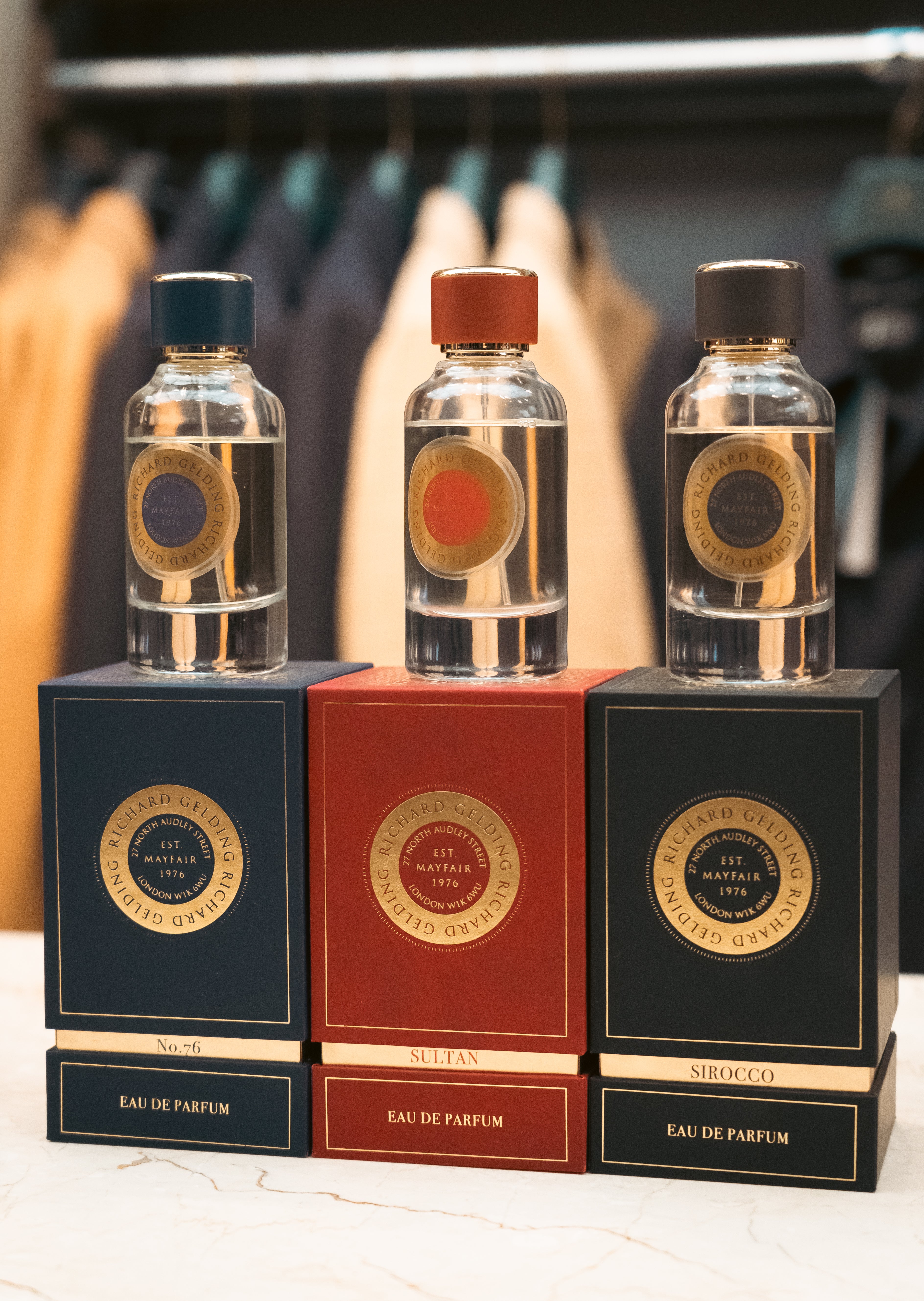 Three glass perfume bottles with colored caps rest on boxes, including Richard Gelding Sirocco EdP 100ml. Blurred coats in the background highlight Sirocco's long-lasting, oriental fragrance by Richard Gelding. , available at Richard Gelding Mayfair