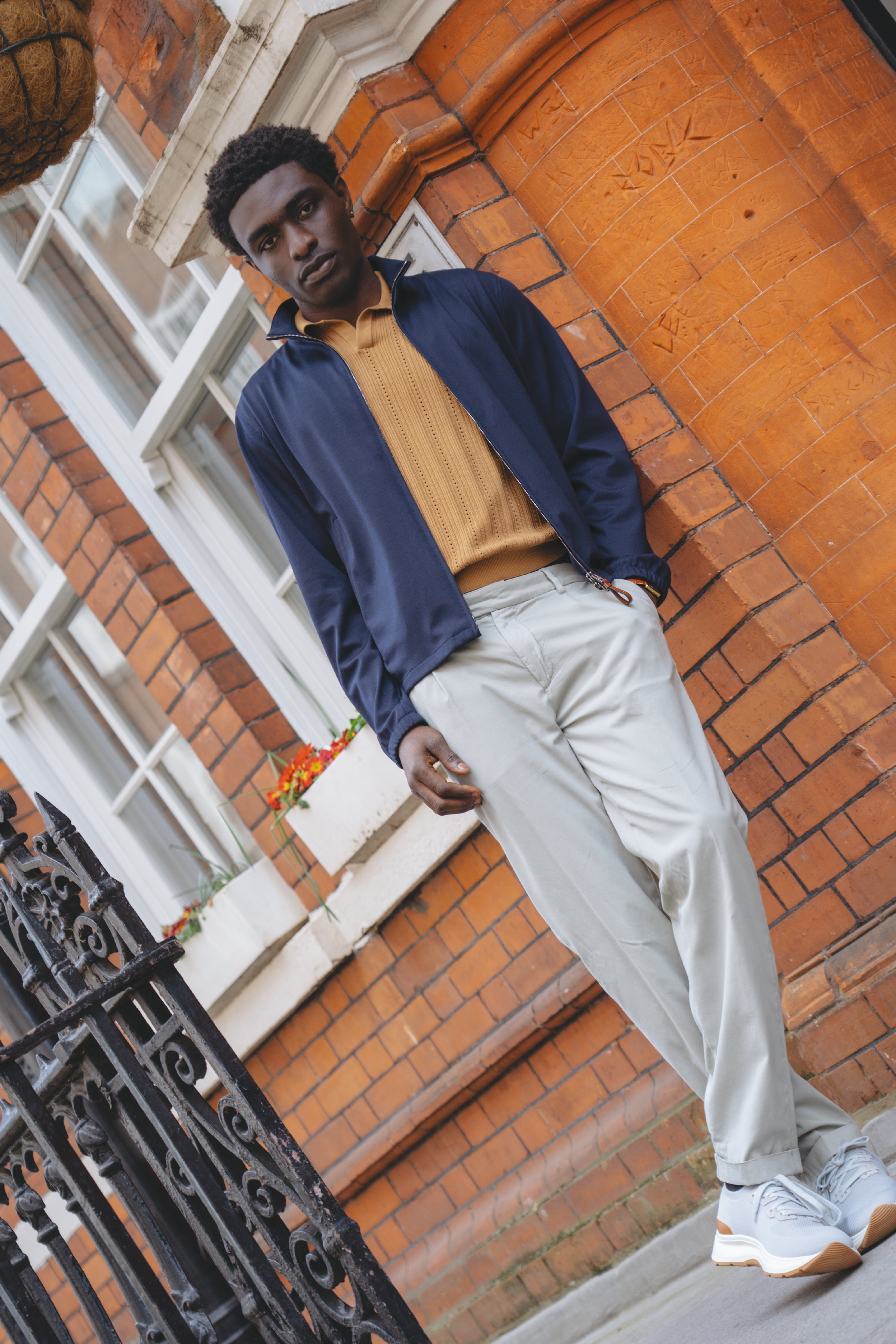 A man stands casually against an orange-brick building, wearing a Manto Navy Loro Piana Wool/Silk Rain System Asiago Blouson, mustard shirt, beige pants, and white sneakers with orange accents. A wrought-iron fence and window flower box are visible. , available at Richard Gelding Mayfair