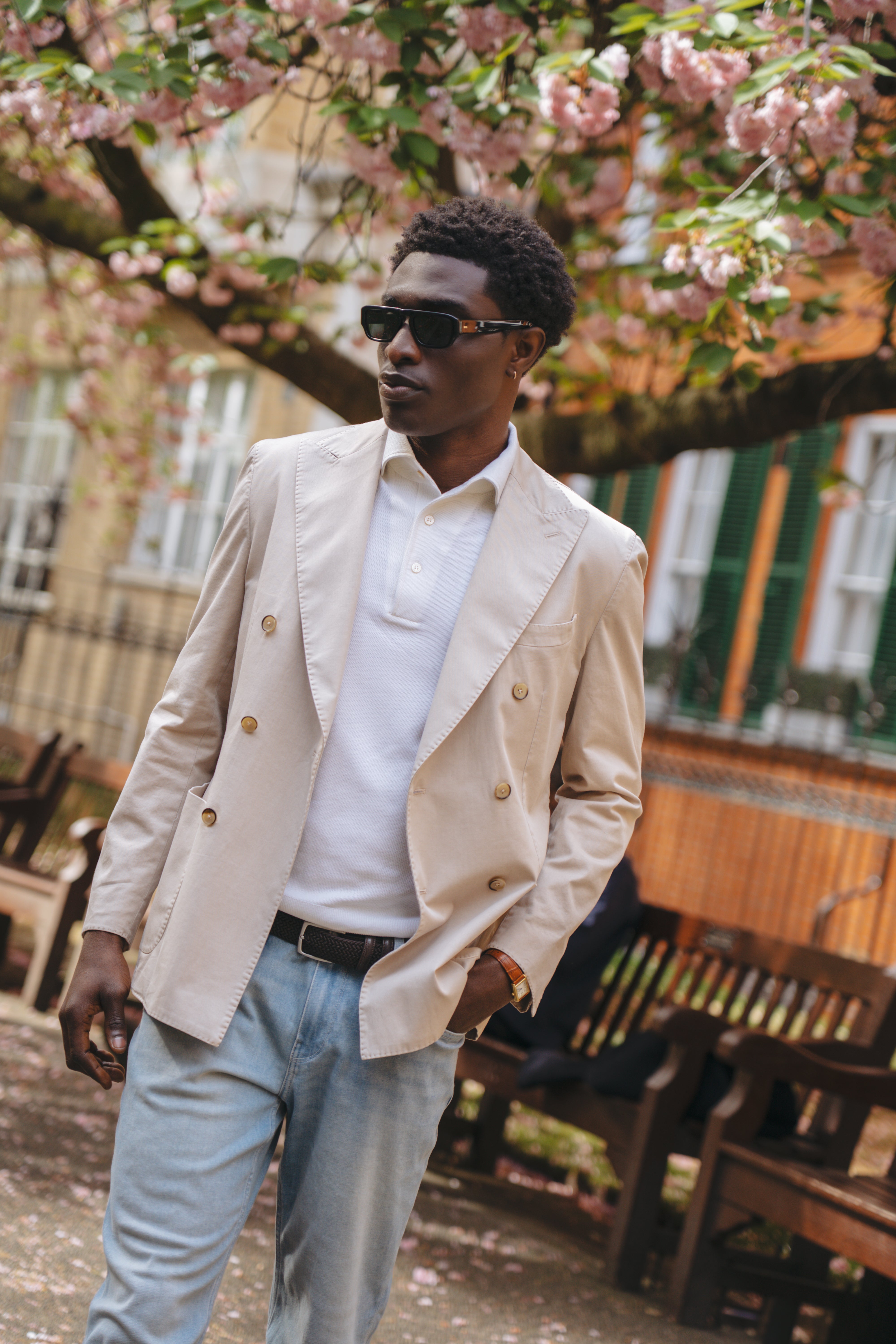 A man wearing sunglasses, a cream double-breasted blazer, a white polo shirt, and Paige Ferdinand Blue Cotton Transcend Normandie Straight Leg Jeans stands outdoors among pink-blossomed trees and wooden benches, evoking a stylish spring atmosphere. , available at Richard Gelding Mayfair