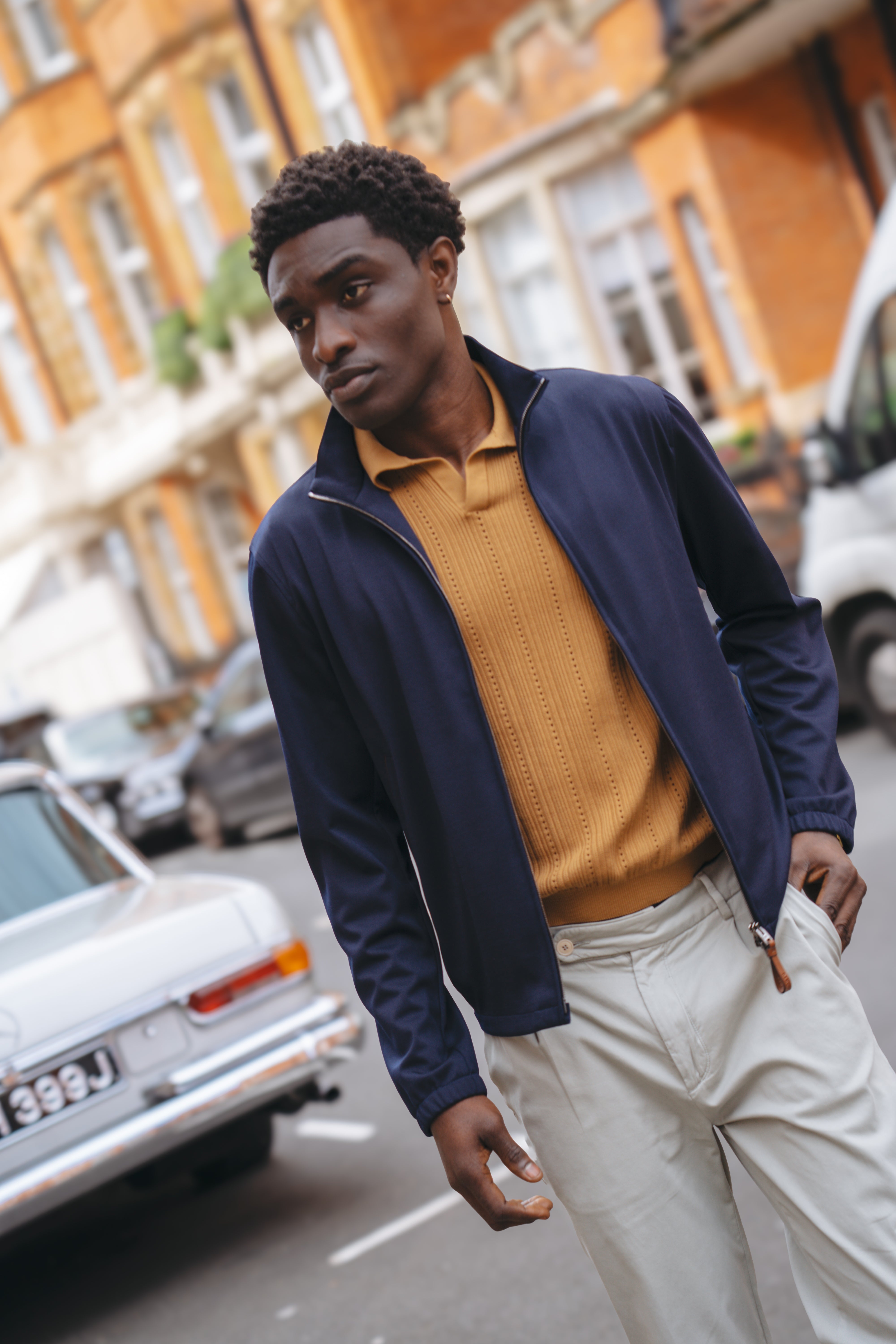 A man with short curly hair and a serious look walks past vintage cars and orange brick buildings, wearing a Manto Navy Loro Piana Wool/Silk Rain System Asiago Blouson over a mustard shirt and cream pants. , available at Richard Gelding Mayfair