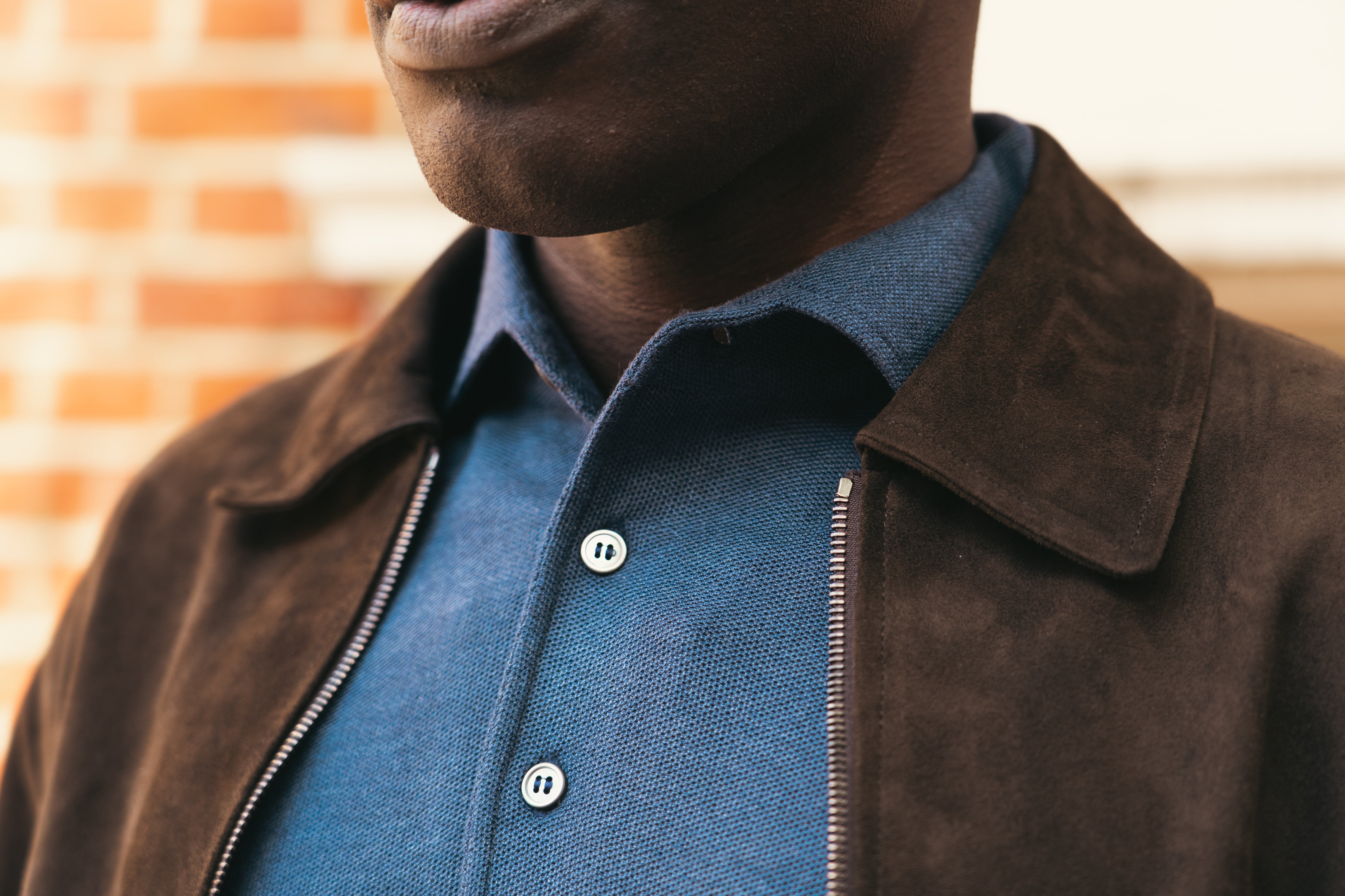 A close-up shows a person wearing the Gran Sasso Navy Blue Silk Pique Polo, with only the lower face, neck, and upper torso visible. The blurred background features a brick wall and a light-colored structure. , available at Richard Gelding Mayfair