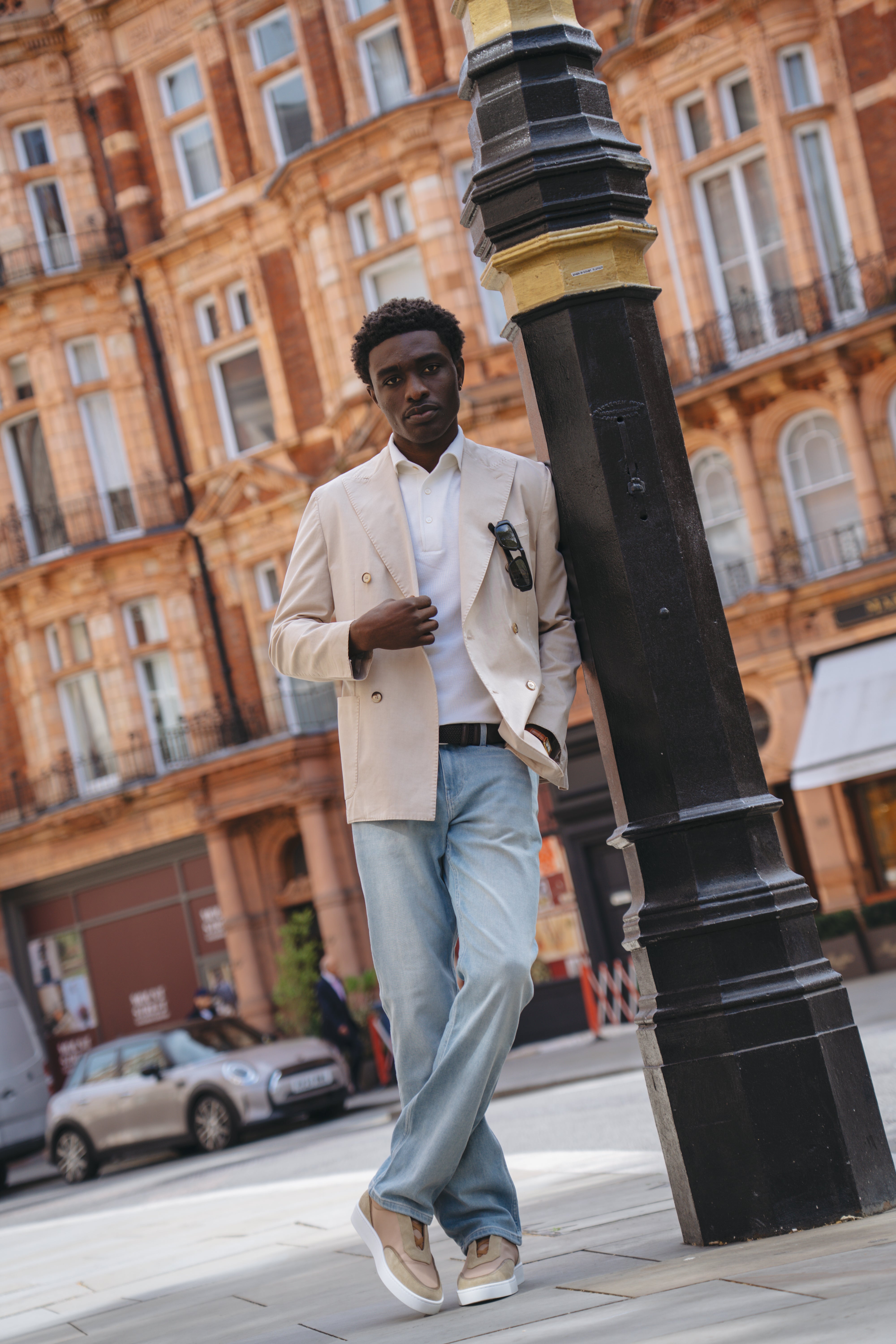 Wearing Paige Ferdinand Blue Cotton Transcend Normandie Straight Leg Jeans, a man in a tan blazer and white shirt leans against a black lamppost on a city street. He holds sunglasses, looking relaxed and stylish amid ornate brick buildings. , available at Richard Gelding Mayfair
