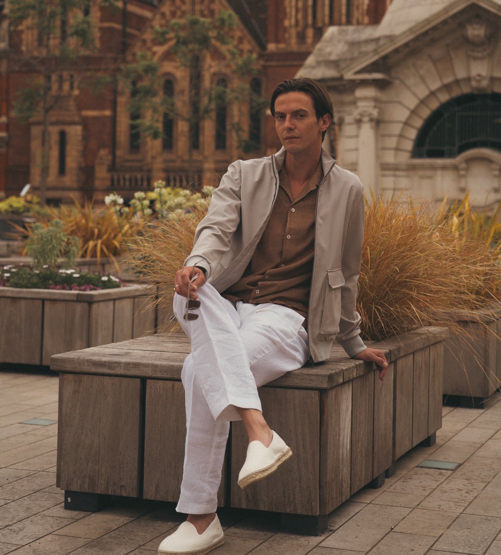 A man with dark hair sits on a wooden bench outdoors, holding sunglasses. He wears a light gray jacket, brown shirt, white pants, and white slip-on shoes. Behind him are planter boxes with yellow grasses and a historic building with arched windows. , available at Richard Gelding Mayfair