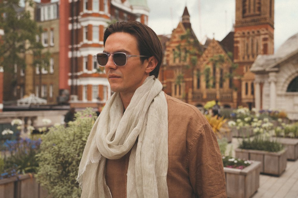 A man wearing sunglasses, a light beige scarf, and a brown shirt stands outdoors in an urban garden. Historical brick buildings with ornate architectural details and greenery are visible in the background under a cloudy sky. , available at Richard Gelding Mayfair