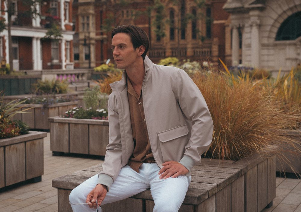 A man with short dark hair sits on a wooden bench in an urban outdoor area. He wears a light gray jacket, brown shirt, and light blue jeans. Ornamental grasses and plants fill raised beds around him, and historic brick buildings are in the background. , available at Richard Gelding Mayfair