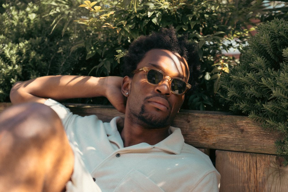 A man wearing sunglasses and a light-colored shirt relaxes outdoors, leaning against a wooden bench surrounded by green leafy bushes. Sunlight and shadows fall across his face and body, giving a calm and tranquil atmosphere. , available at Richard Gelding Mayfair
