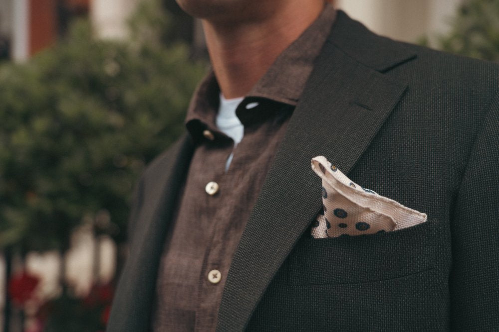 A man in a textured dark suit jacket and brown buttoned shirt stands outdoors. A beige pocket square with dark polka dots is neatly folded in his breast pocket. Blurred greenery is visible in the background. , available at Richard Gelding Mayfair