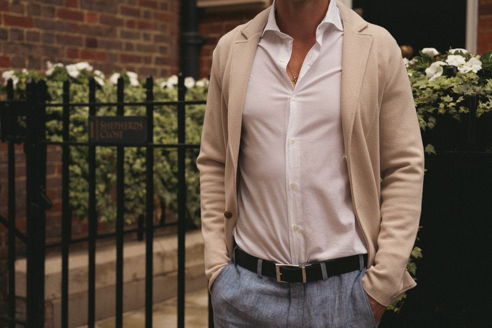 A man stands outdoors by a black iron fence and white flowers, wearing a beige blazer over an unbuttoned white shirt and light blue trousers. His hands are in his pockets and his face is out of frame. The background includes brick walls and steps. , available at Richard Gelding Mayfair