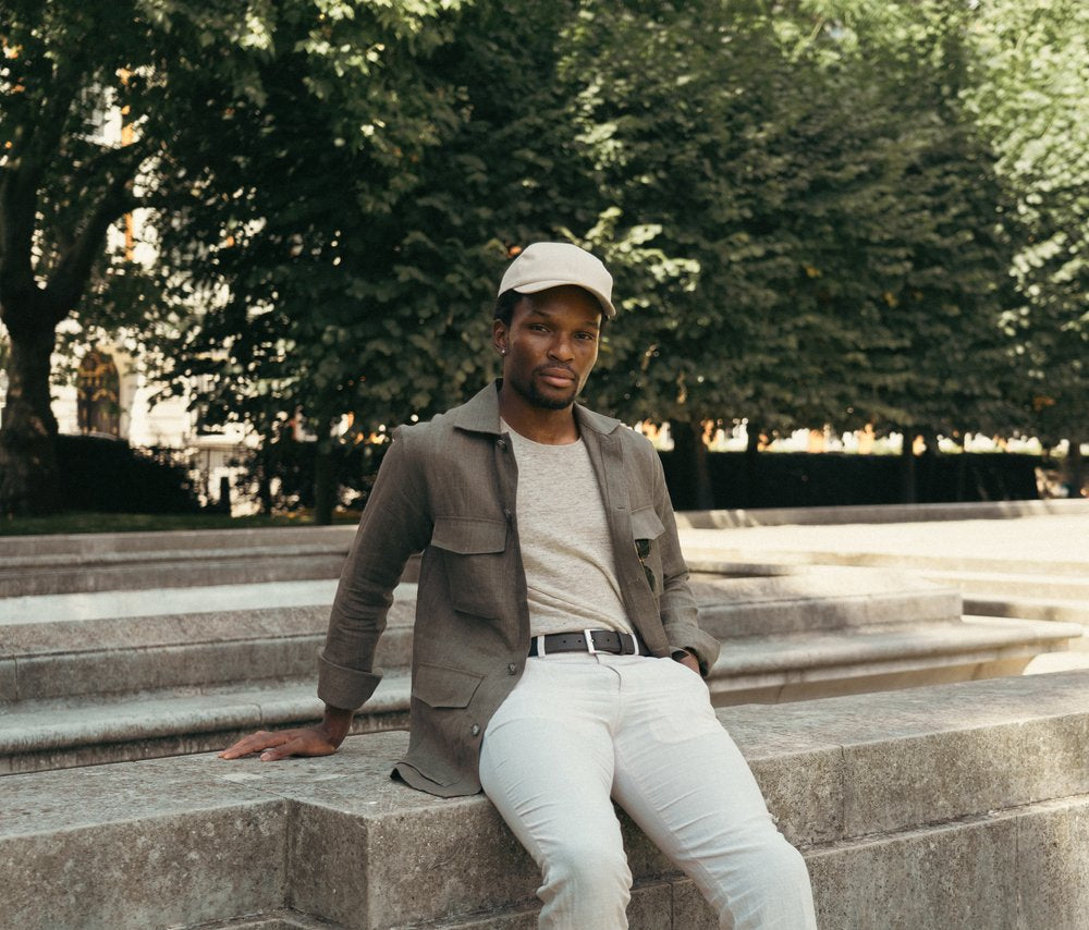 A man in light-colored pants, a gray t-shirt, olive jacket, and beige cap sits on a concrete bench outdoors. He looks at the camera with a relaxed expression. Behind him are large, leafy green trees and dappled sunlight. , available at Richard Gelding Mayfair