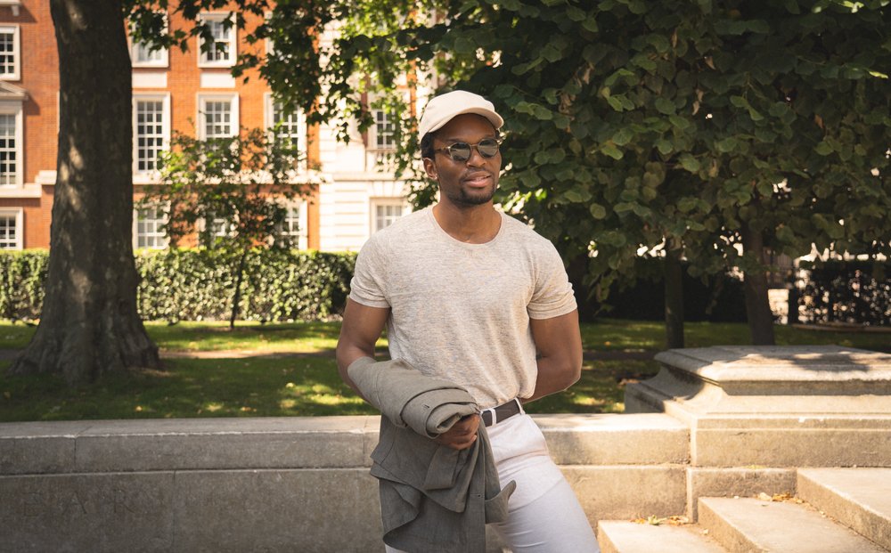 A man wearing a light beige cap, sunglasses, a gray t-shirt, and white pants stands outdoors, holding a gray jacket. He is in a sunny park with green grass, trees, and a red-brick building with white-trimmed windows in the background. , available at Richard Gelding Mayfair