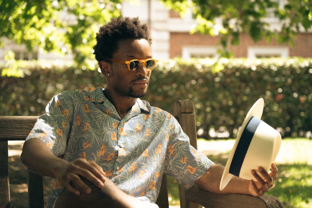A man wearing yellow sunglasses and a floral shirt sits on a wooden bench outdoors, holding a straw fedora hat with a blue band. Sunlight filters through trees, casting dappled shadows. Bushes and a brick building are blurred in the background. , available at Richard Gelding Mayfair