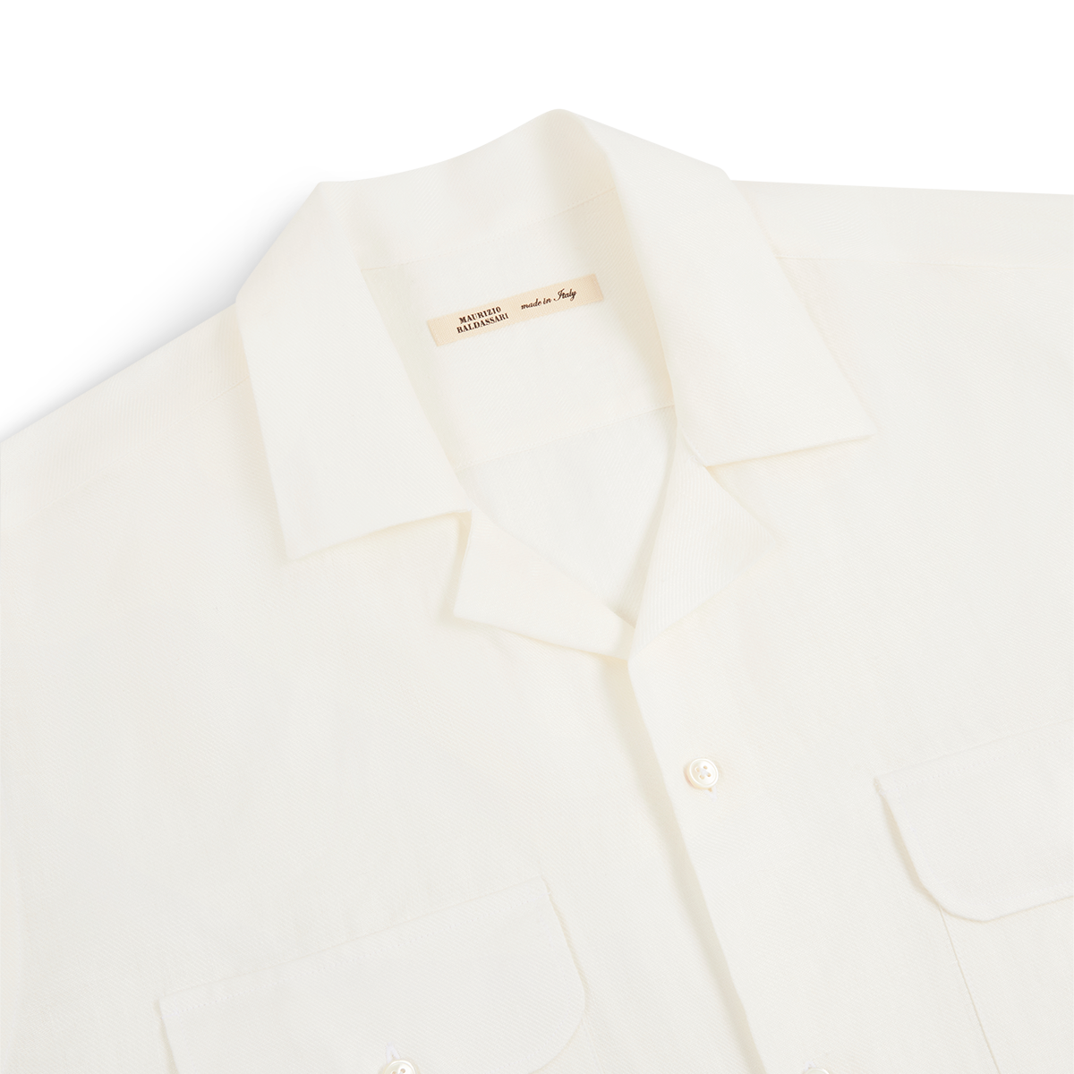 A close-up of the Maurizio Baldassari White Linen Army Shirt shows its short sleeves, notched collar, two buttoned chest pockets, and lightweight linen fabric. A beige inner tag displays the product name. The shirt is set against a plain white background. , available at Richard Gelding Mayfair