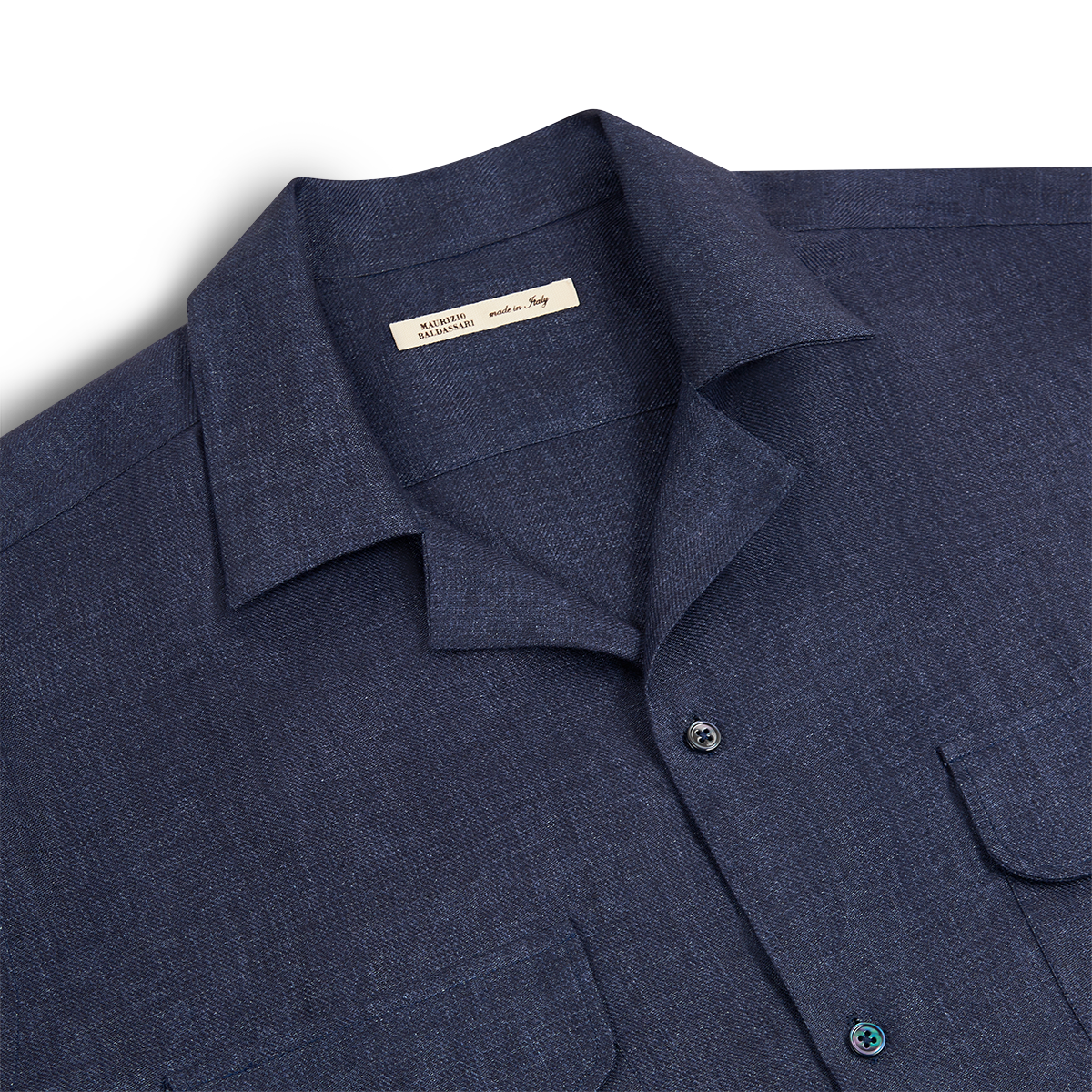 The Maurizio Baldassari Denim Blue Linen Army Shirt, featuring a textured fabric, two chest pockets, and black buttons, is neatly folded with its inner tag visible and laid flat on a white background. , available at Richard Gelding Mayfair