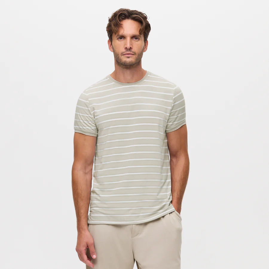 A man with wavy brown hair wears the Homebody Beige Desert Stripe Modal Marlow Striped T-Shirt, featuring thin white stripes, paired with beige pants. He stands facing the camera with one hand in his pocket against a white background. , available at Richard Gelding Mayfair