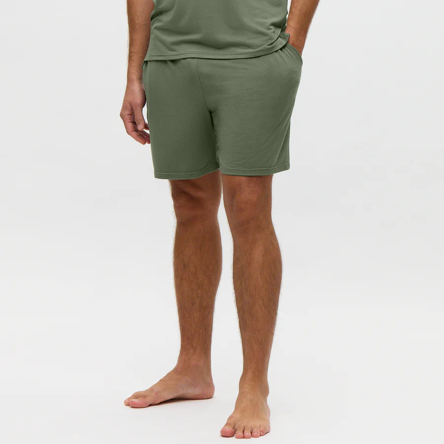 A person stands barefoot on a plain light background, hands in their pockets, wearing Homebody’s Charred Green Modal Wimbledon Shorts. The relaxed fit shorts are the focus, with the image cropped at shoulders and feet. , available at Richard Gelding Mayfair