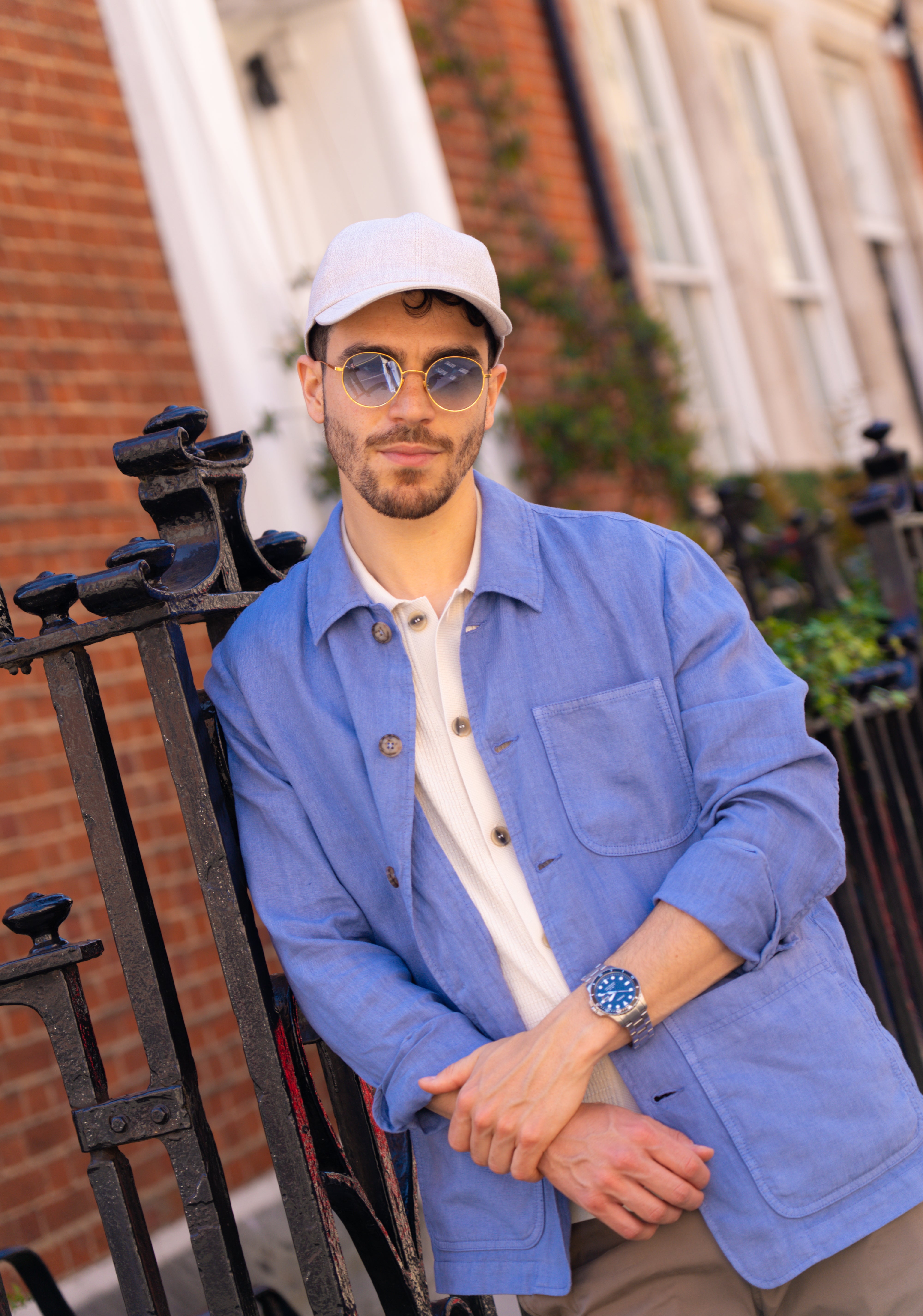 A man with a trimmed beard and mustache leans on a black iron fence, wearing an Altea Sugar Packet Blue Linen Chore Jacket, a light gray cap, round sunglasses, and a pale shirt. Red brick buildings and greenery form the backdrop. , available at Richard Gelding Mayfair
