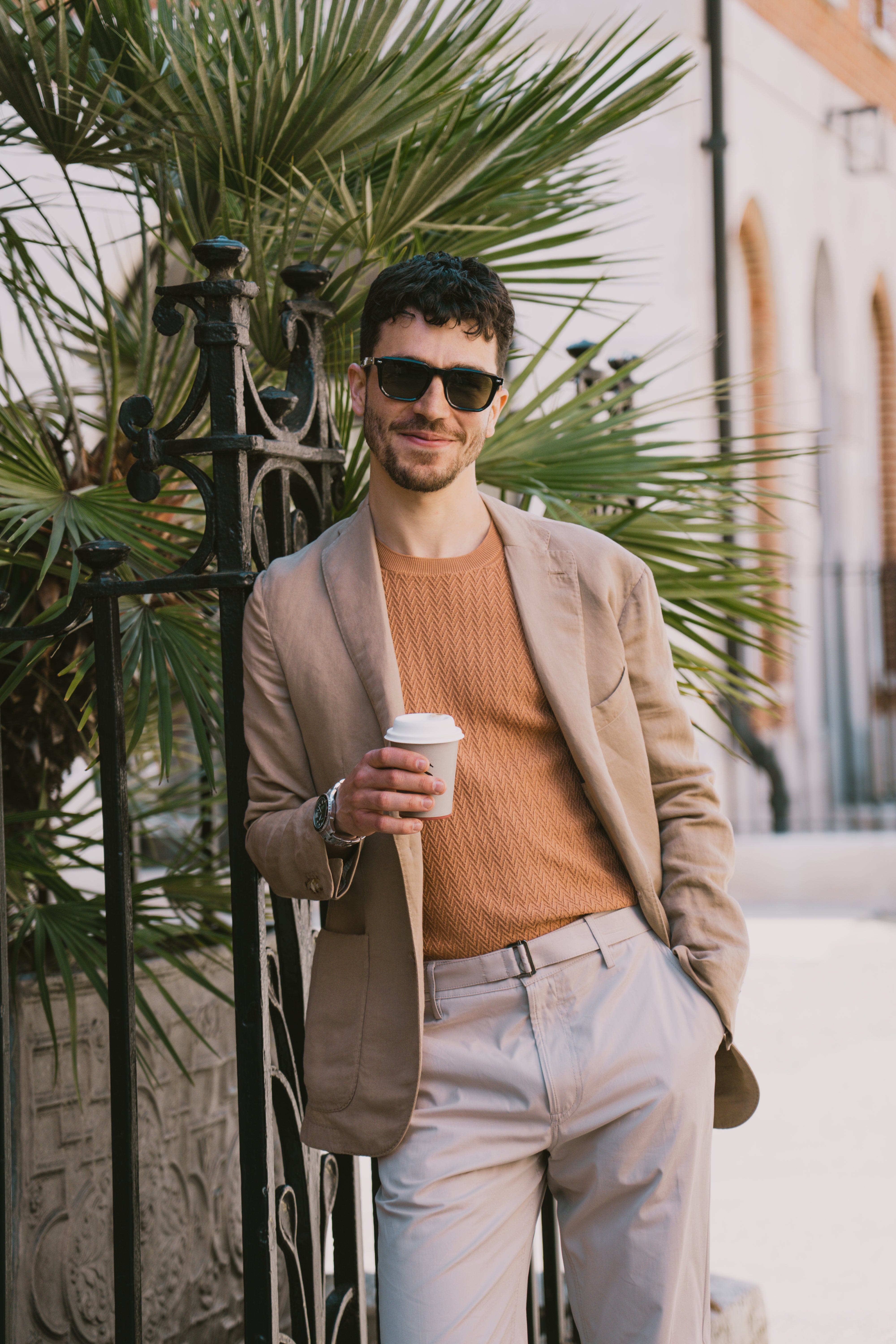 A man in a Boglioli Camel Beige Satin Cotton/Linen Jacket leans against a black iron fence, holding a white coffee cup, with a large green palm and arched windows in the background. He's also wearing sunglasses and light beige trousers. , available at Richard Gelding Mayfair
