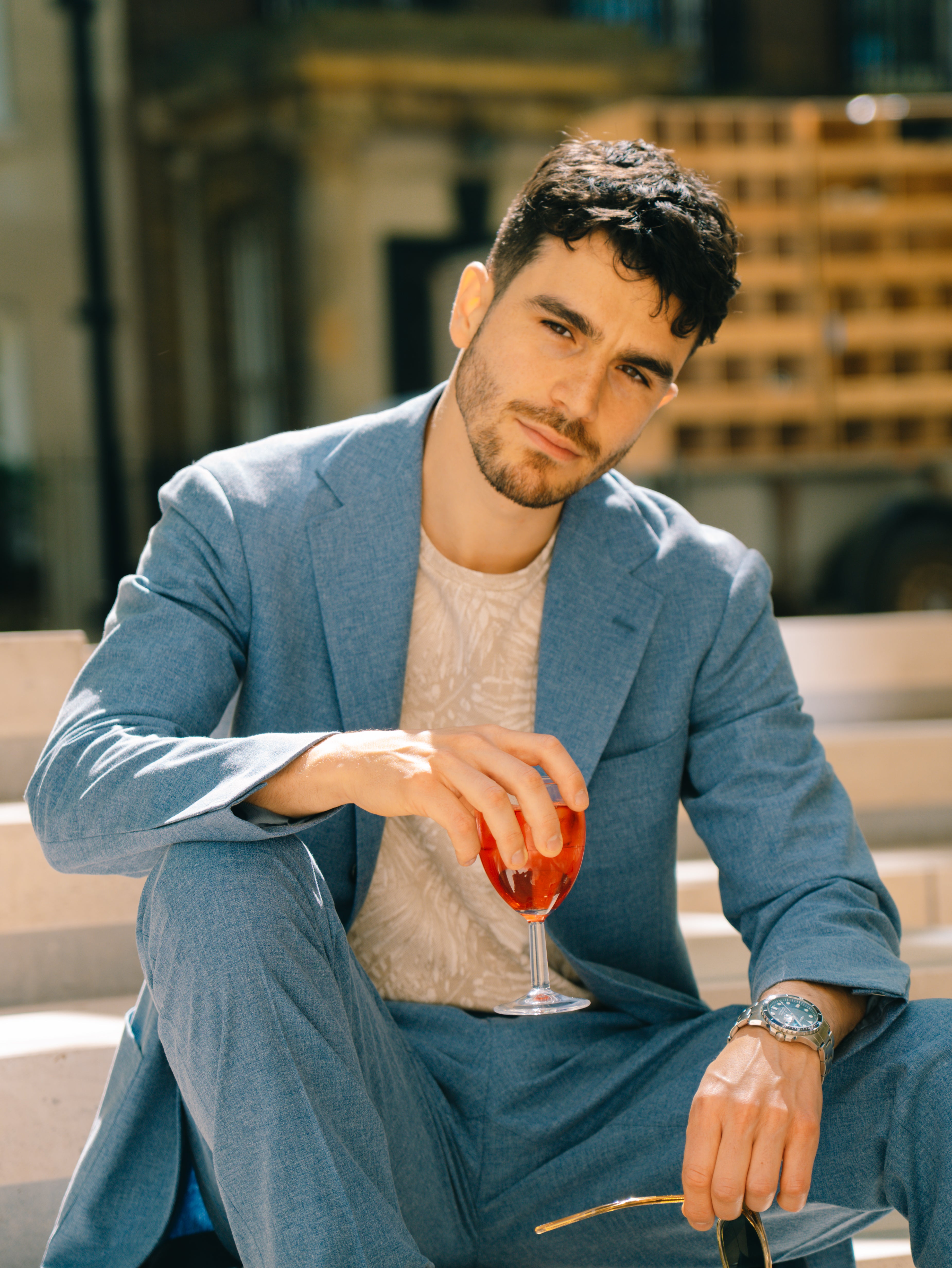 A man with short dark hair and a trimmed beard sits outdoors in sunlight, wearing the Ring Jacket Pale Blue Melange Tropical Wool/Cotton/Silk Suit over a patterned beige shirt. He holds sunglasses and a glass of red drink, looking calm. Blurred steps and a building are behind him. , available at Richard Gelding Mayfair