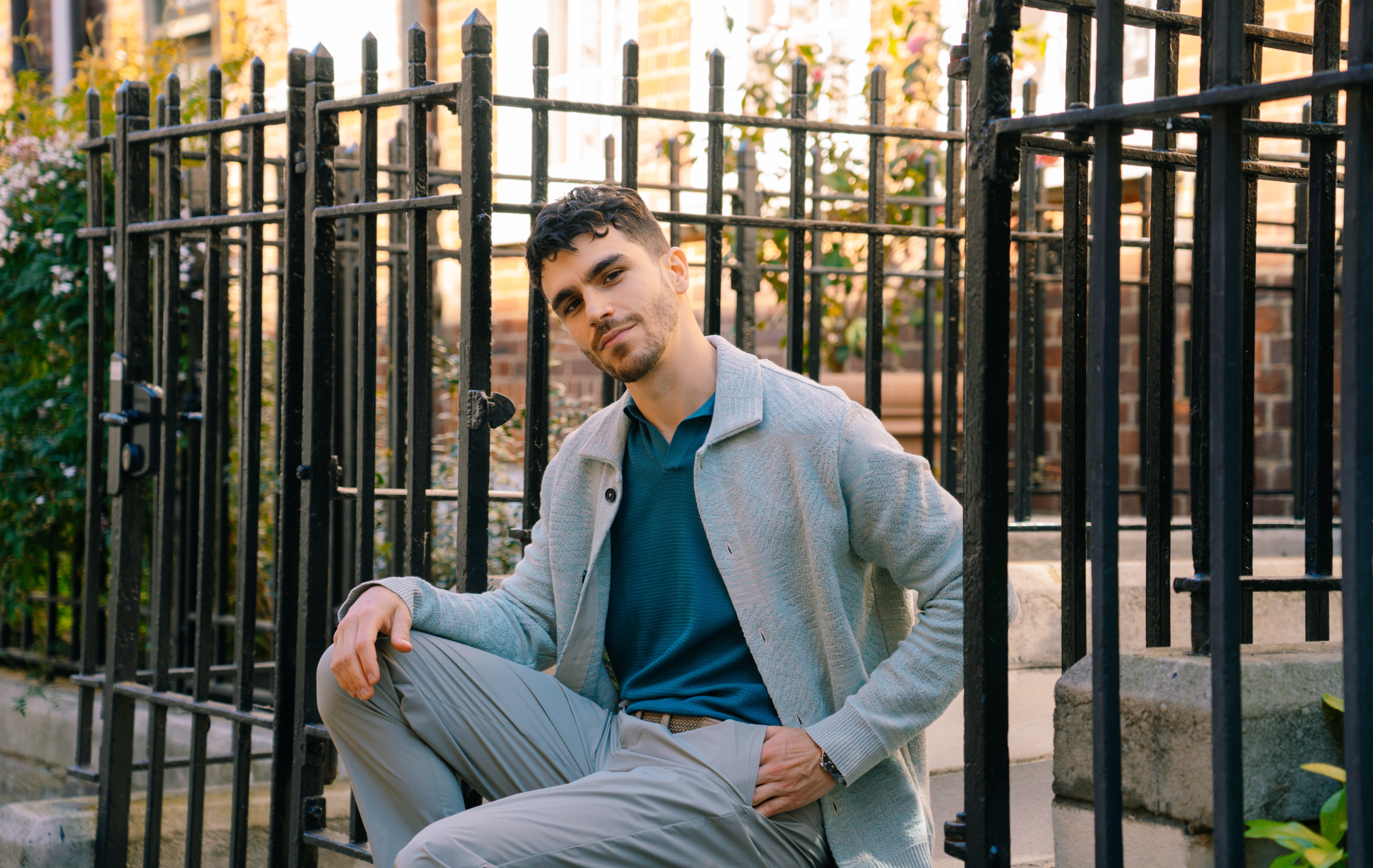 A man with short, dark curly hair and a trimmed beard sits on outdoor stone steps in a Maurizio Baldassari Aqua Green Cotton/Linen Blend Jacquard Palma Overshirt, teal shirt, and matching pants. Black railings and plants are in the background. , available at Richard Gelding Mayfair