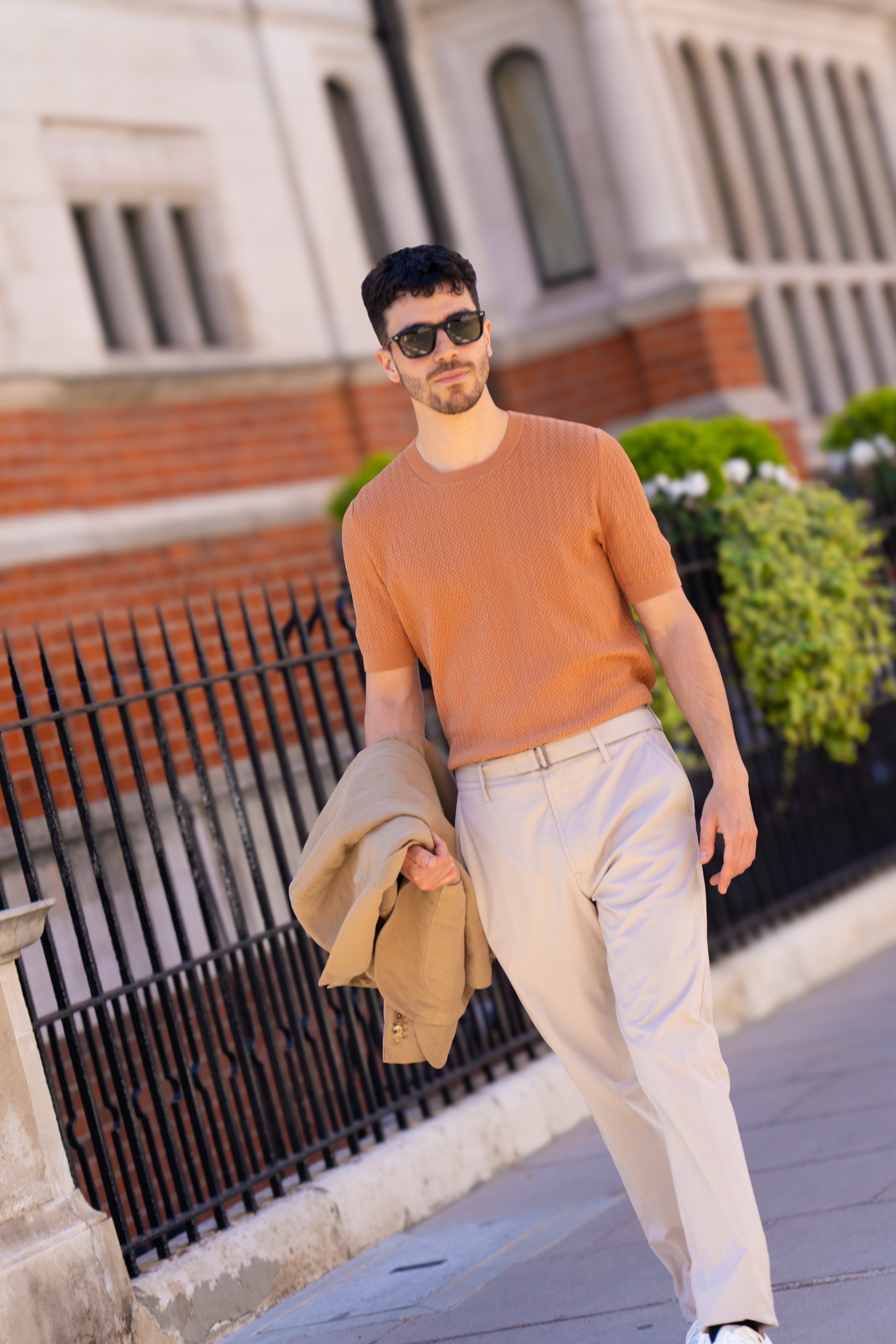 A man with short dark hair and sunglasses walks on a city sidewalk wearing a Gran Sasso Rust Chevron Weave T-Shirt, beige trousers with a belt, and white sneakers. He carries a tan jacket. A black iron fence and greenery are behind him. , available at Richard Gelding Mayfair