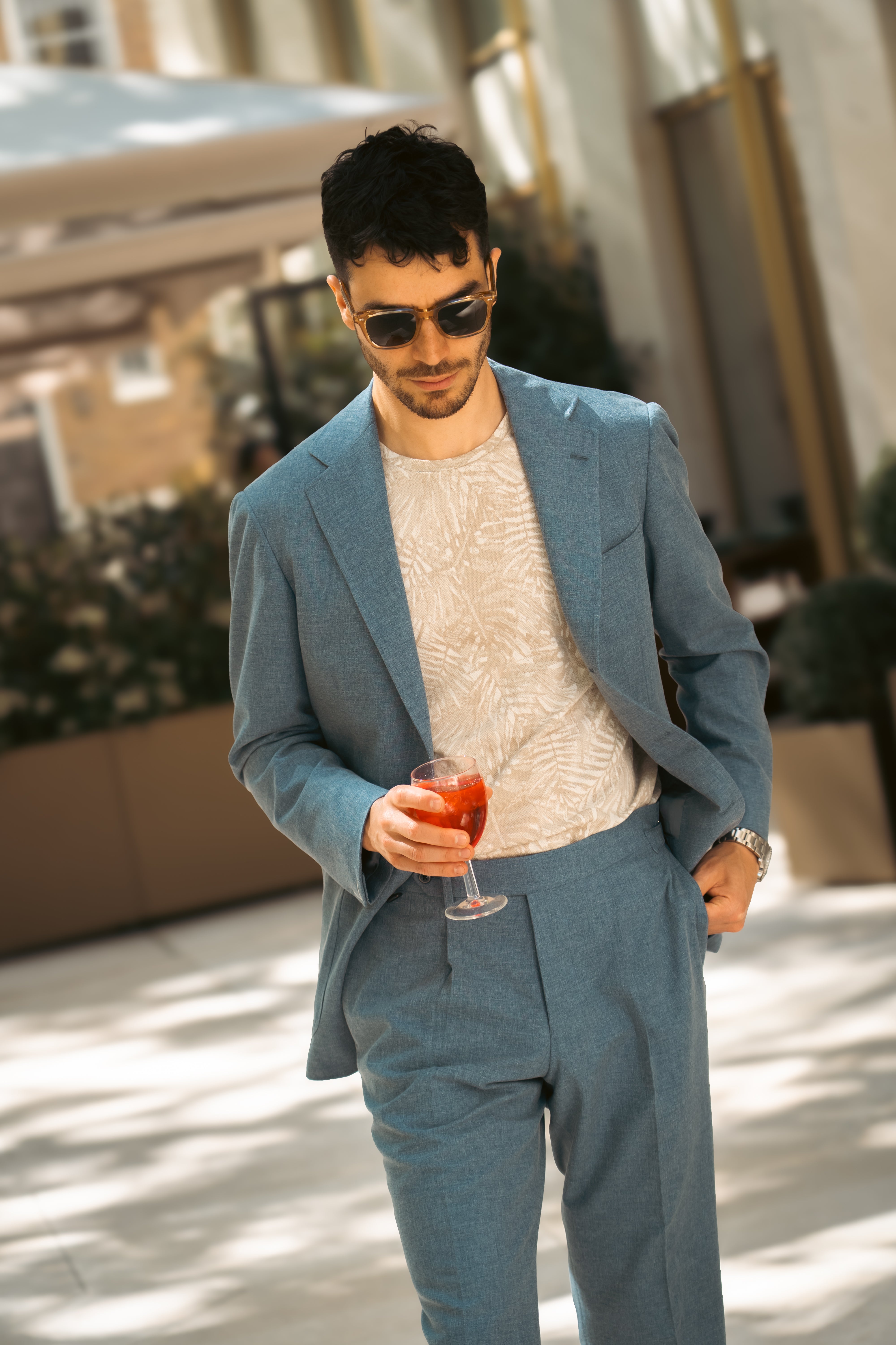 A man in a light blue suit, Maurizio Baldassari Beige Silk/Cotton Palm Tree Print T-Shirt, and sunglasses walks outdoors with a red cocktail in a wine glass. He has dark hair and facial hair; blurred plants and modern buildings are behind him. , available at Richard Gelding Mayfair