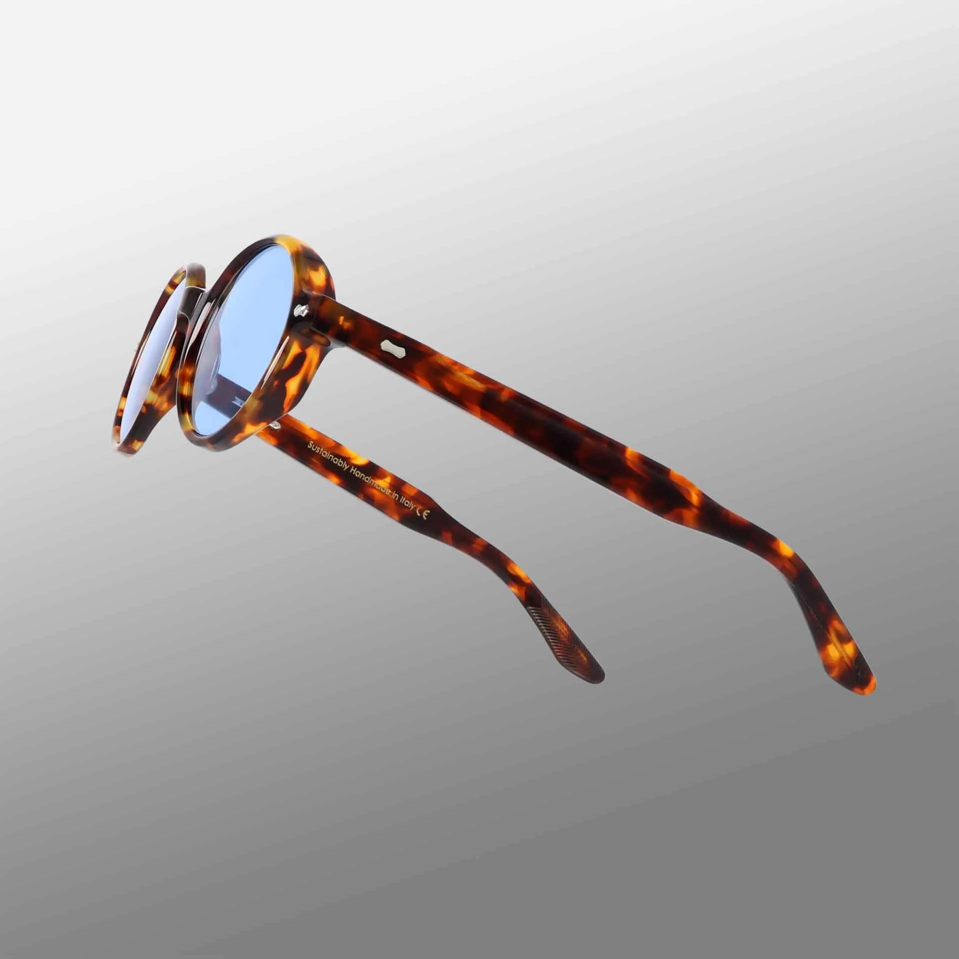 The Giotto Eco Spotted Havana | Blue sunglasses by TBD Eyewear feature blue lenses and tortoiseshell-patterned frames and arms, shown angled on a white-to-gray gradient background with open arms highlighting the spotted amber and brown design. , available at Richard Gelding Mayfair