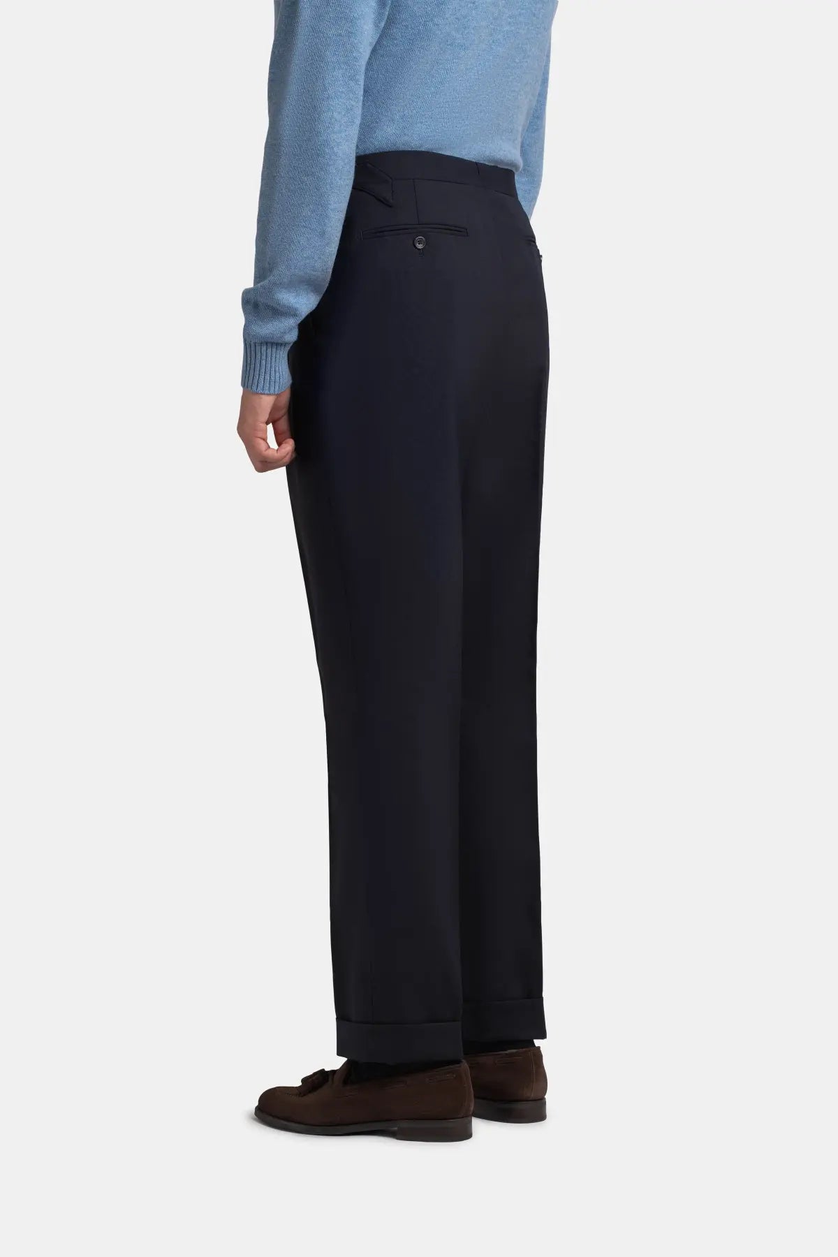A person stands in profile wearing Navy Blue Wide Leg Pleated Twist Wool Trousers by Cavour, featuring a tailored fit and cuffed hems, paired with a light blue long-sleeved sweater and dark brown suede loafers against a plain light background. , available at Richard Gelding Mayfair