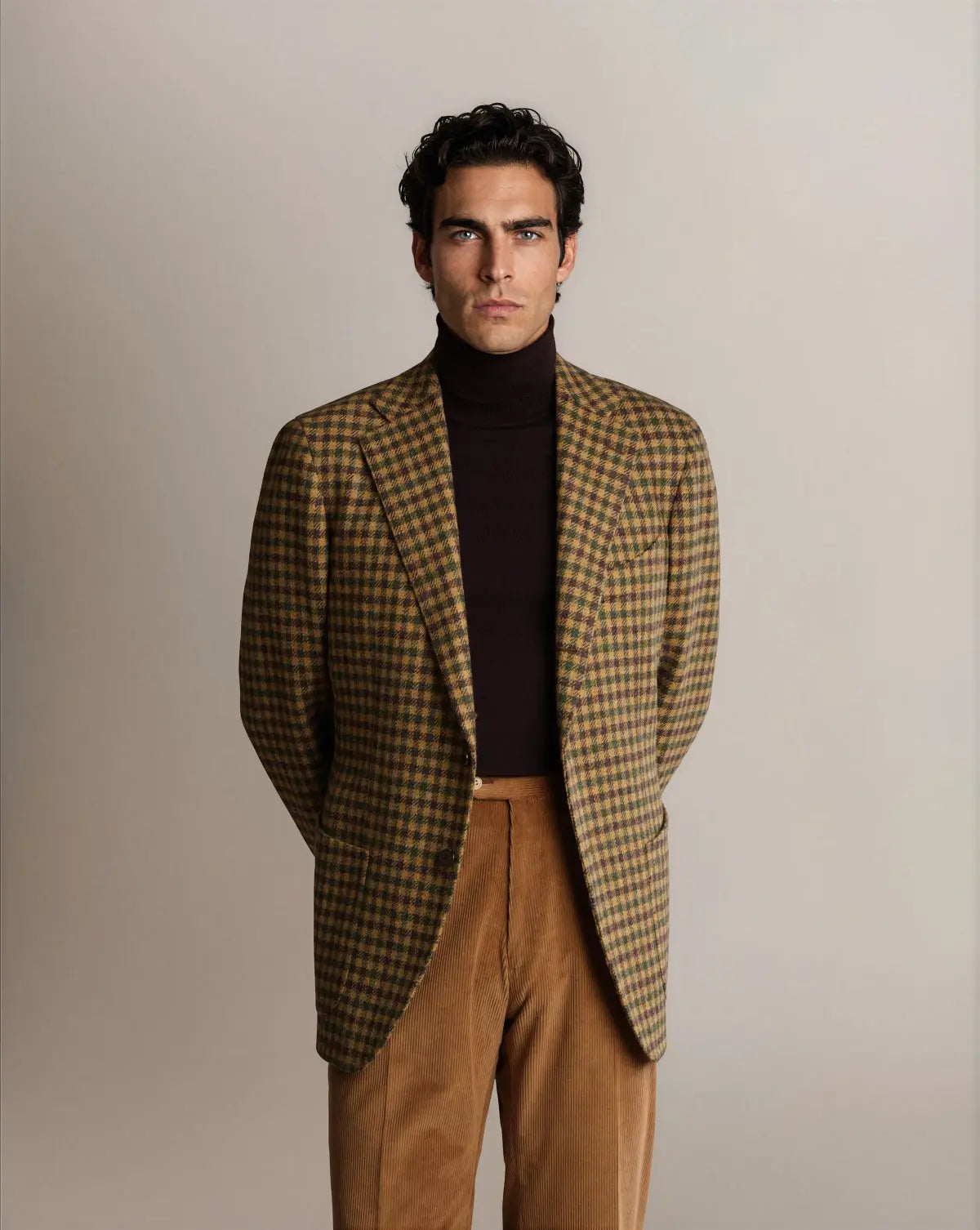 A man with dark hair looks at the camera, wearing a brown and olive Cavour Brown Shepherds Check Tweed Blazer over a dark brown turtleneck and tan corduroy trousers, against a plain light gray studio background. , available at Richard Gelding Mayfair
